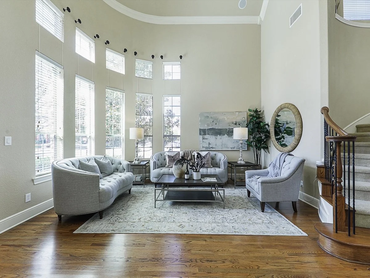 Living room with large floor-to-ceiling windows, white sofas, a coffee table, a rug, side tables with lamps, wall art, a round mirror, a potted plant, and a staircase with a wooden banister.