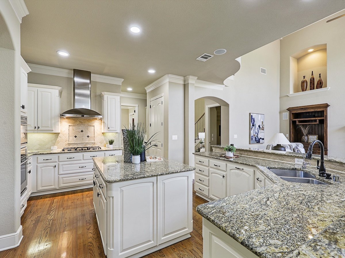 Modern kitchen with white cabinets, granite countertops, a central island with a potted plant, and stainless steel appliances including a range hood. Wood flooring and a living area with decorative items are visible.