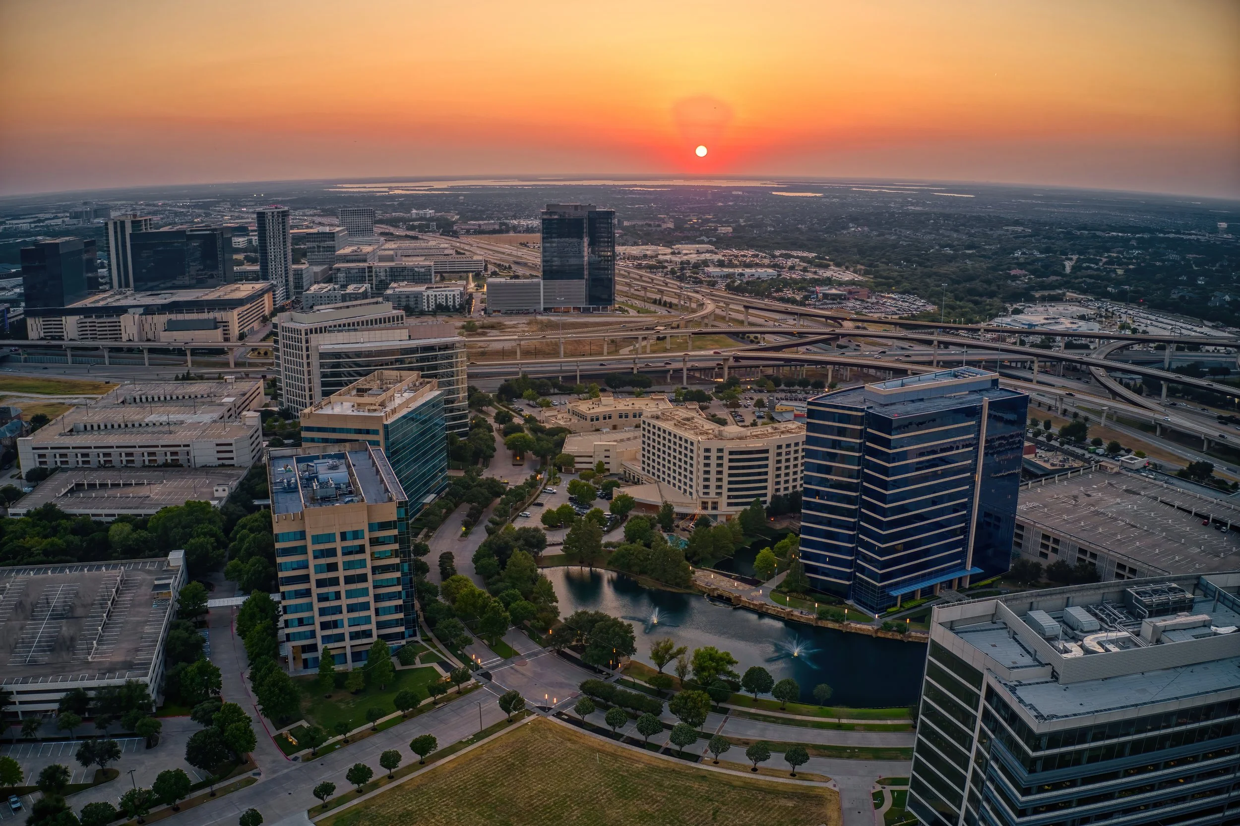 Aerial view of a cityscape at sunset with skyscrapers, a pond with fountains, and a network of highways.