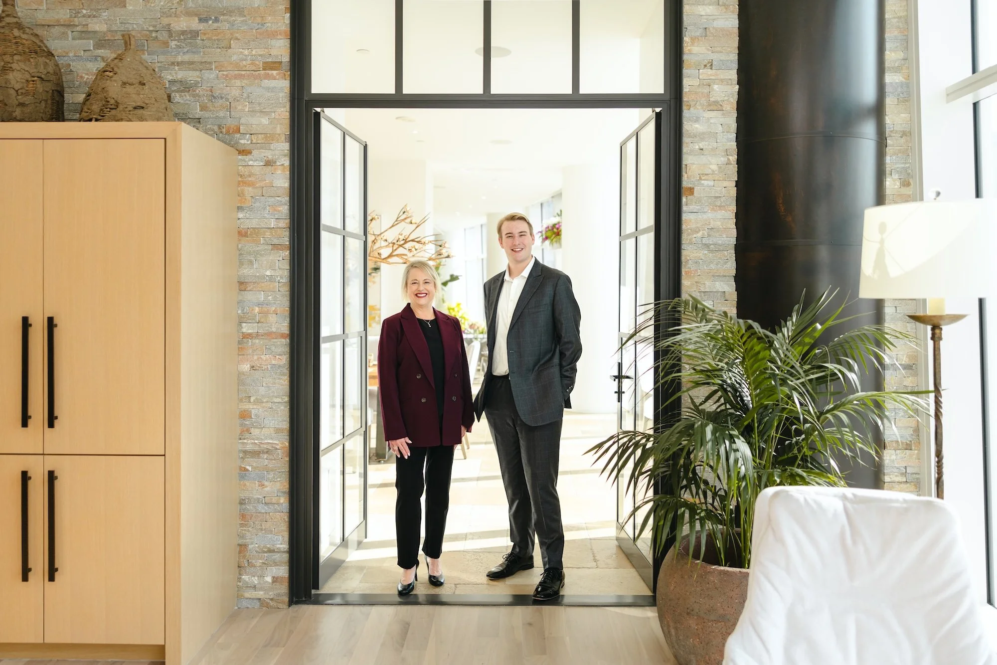Two people, a woman and a man, standing in the doorway of a modern interior space, smiling. The woman has blonde hair and is wearing a burgundy blazer, and the man has short blonde hair and is dressed in a gray suit. The space has a large potted plant, a wooden cabinet, a standing lamp, and a brick wall, with bright natural light coming through the glass doors.