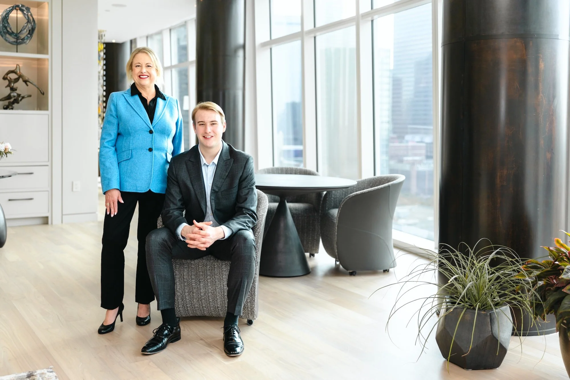 A woman in a blue blazer standing next to a young man in a gray suit sitting in a modern office with large windows, cityscape view, and indoor plants.