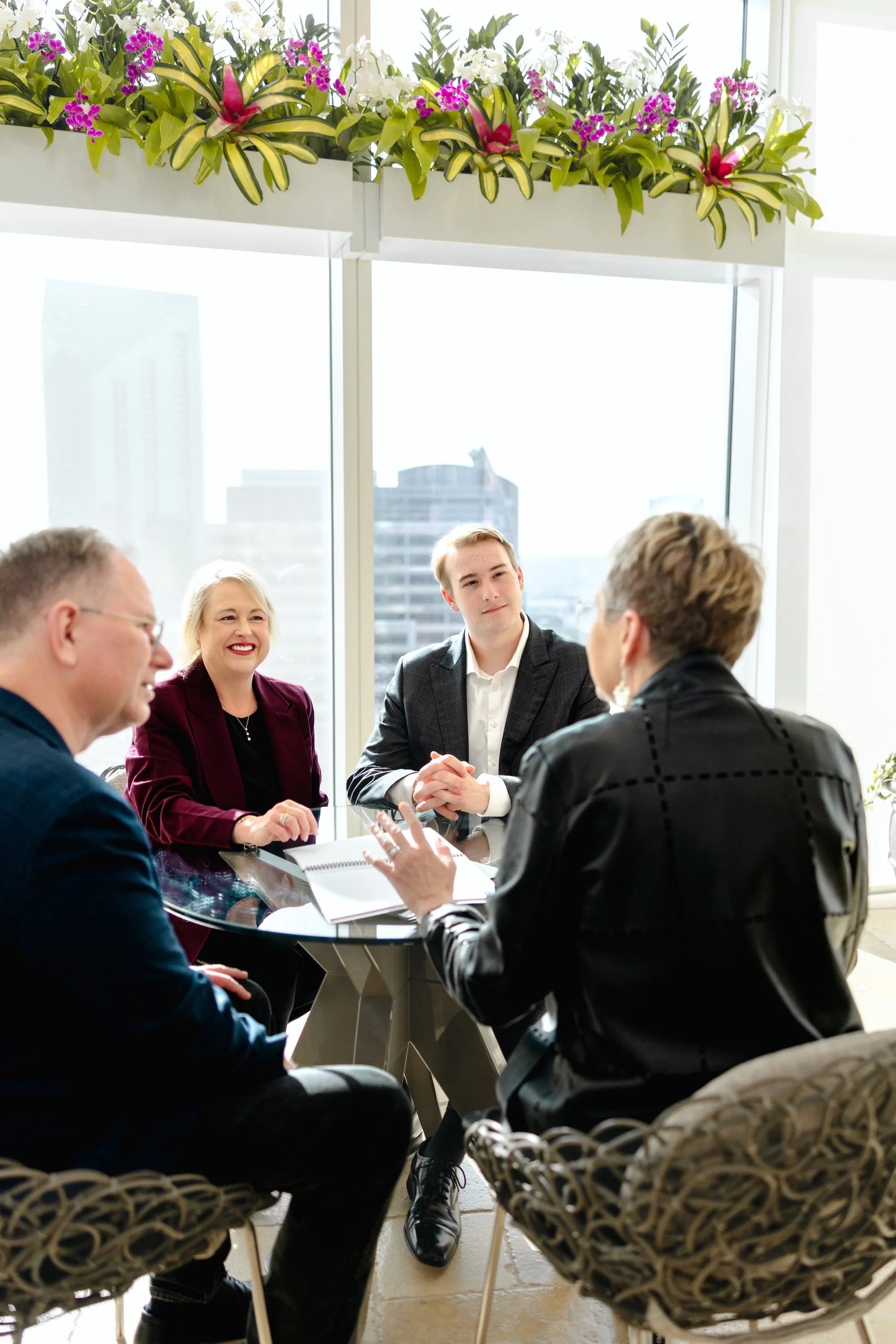 Four professionals in a business meeting in a bright office with large windows and cityscape background, decorated with floral plants on overhead shelf.