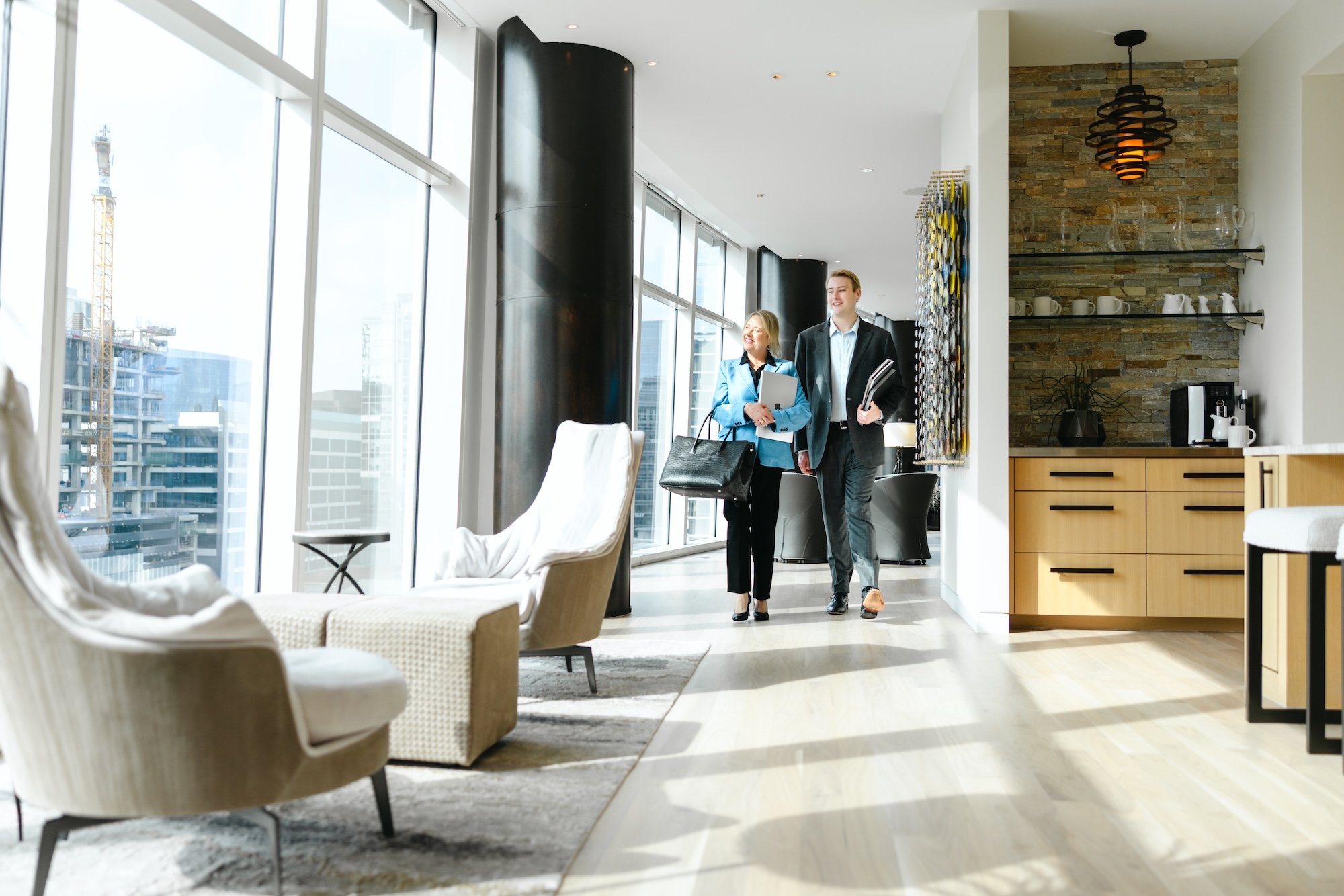 Two business professionals, a woman and a man, walking and talking in a modern office lounge area with large windows, seating, and a kitchenette in the background.