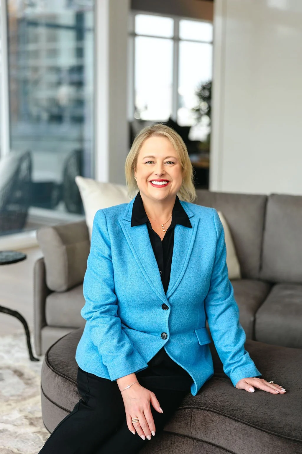 A smiling middle-aged woman with blonde hair wearing a dark purple cardigan sitting at a wooden table with a laptop and a small plant, in a cozy indoor setting.