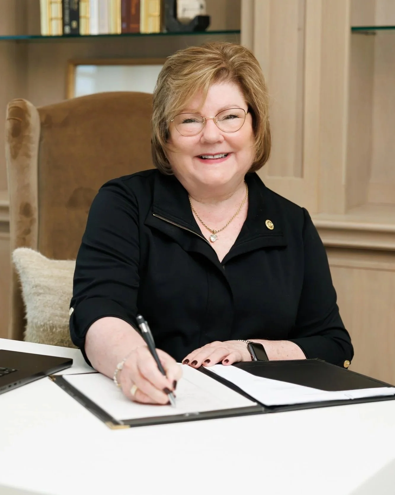 A smiling woman with glasses and short reddish hair sitting at a white table, wearing a black top, gold jewelry, and a smartwatch in a warm-toned room with wooden furnishings.