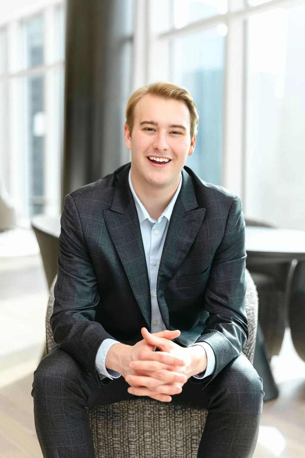 Young man in a black suit and white shirt smiling outdoors with greenery and a building in the background.