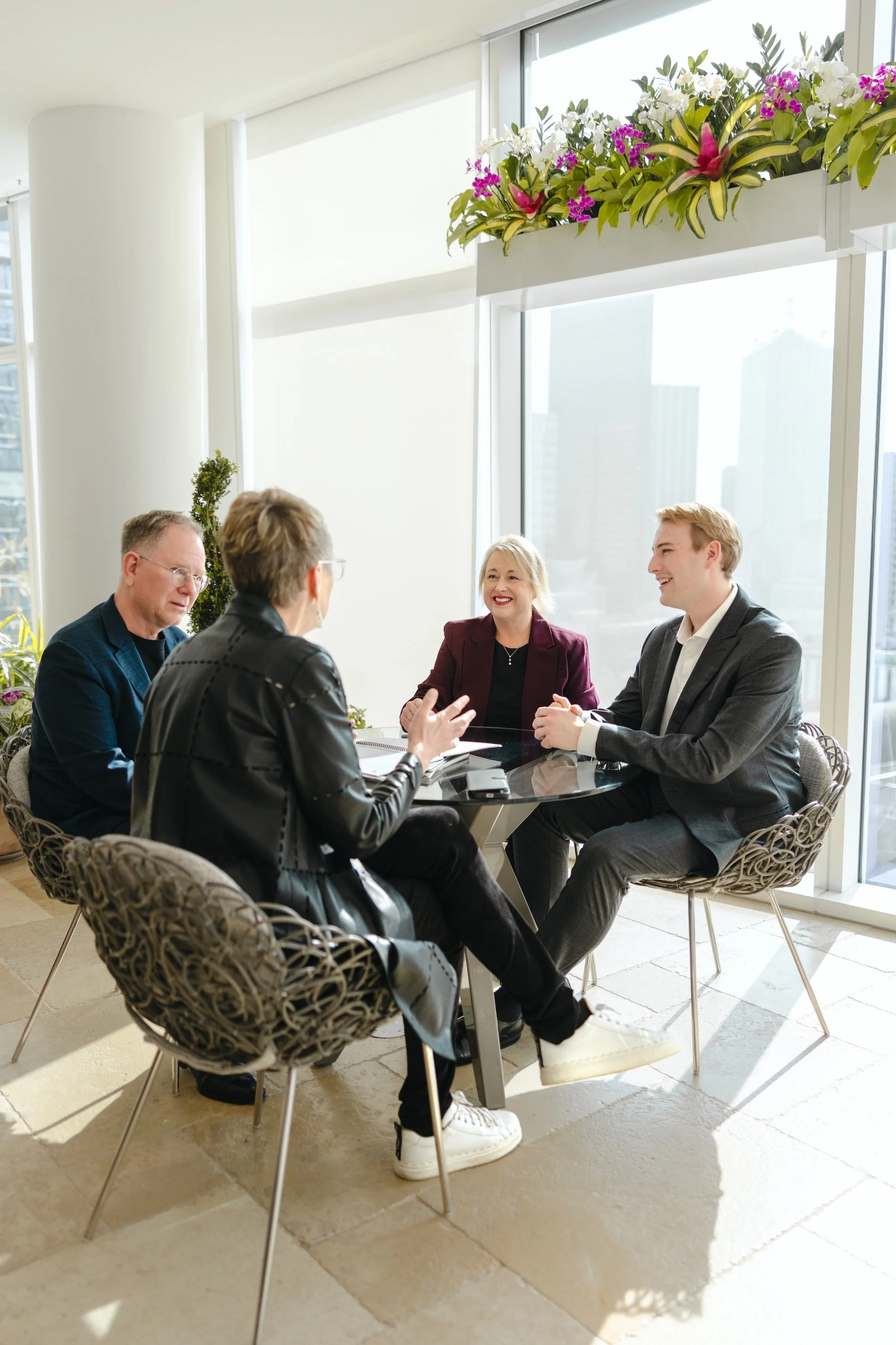 Four people sitting around a glass table in a modern office, engaged in a discussion, with large windows and city buildings in the background.