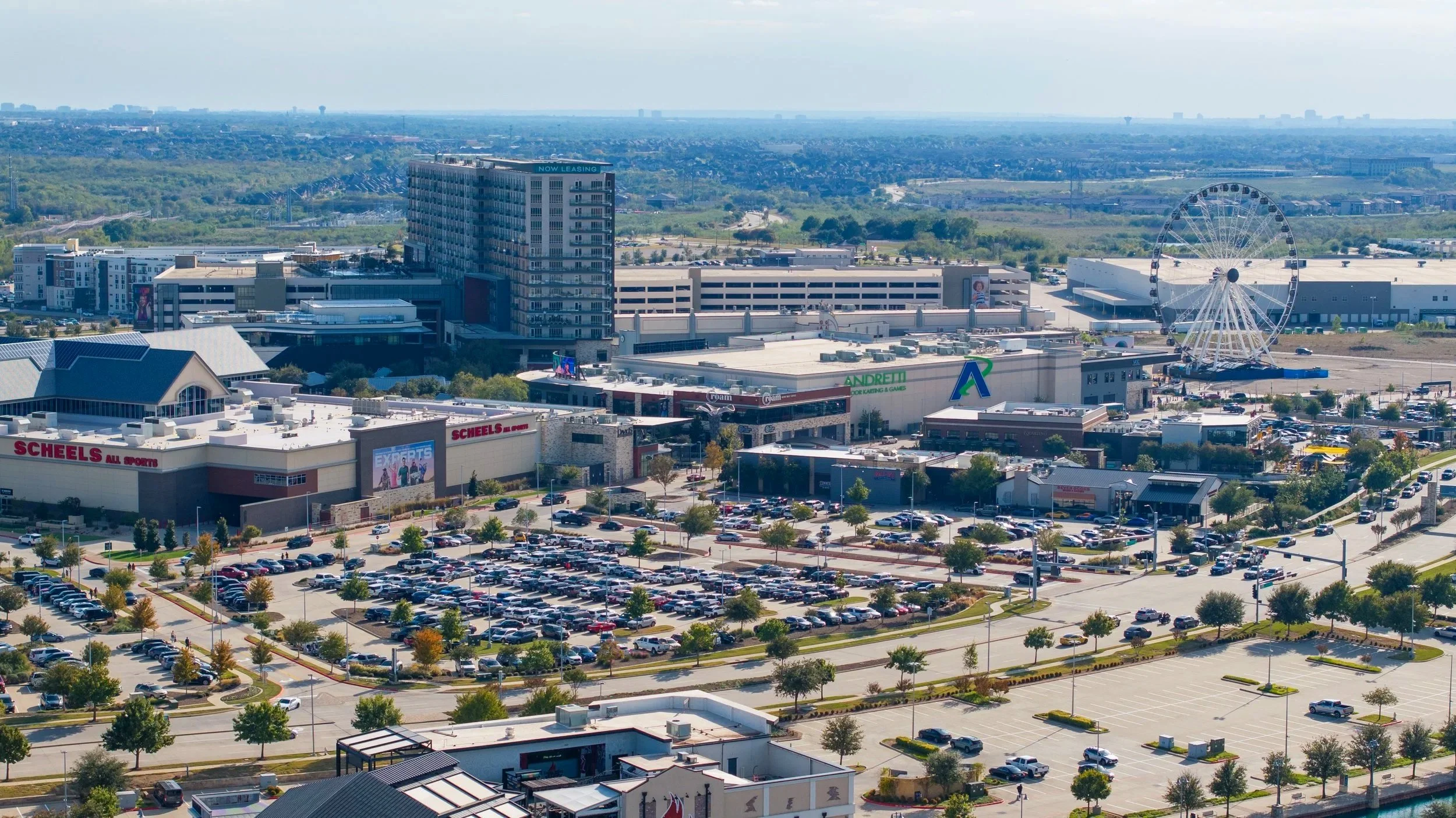 Aerial view of a shopping mall parking lot with many cars, trees, and surrounding buildings, including a Ferris wheel and a tall hotel in the background.