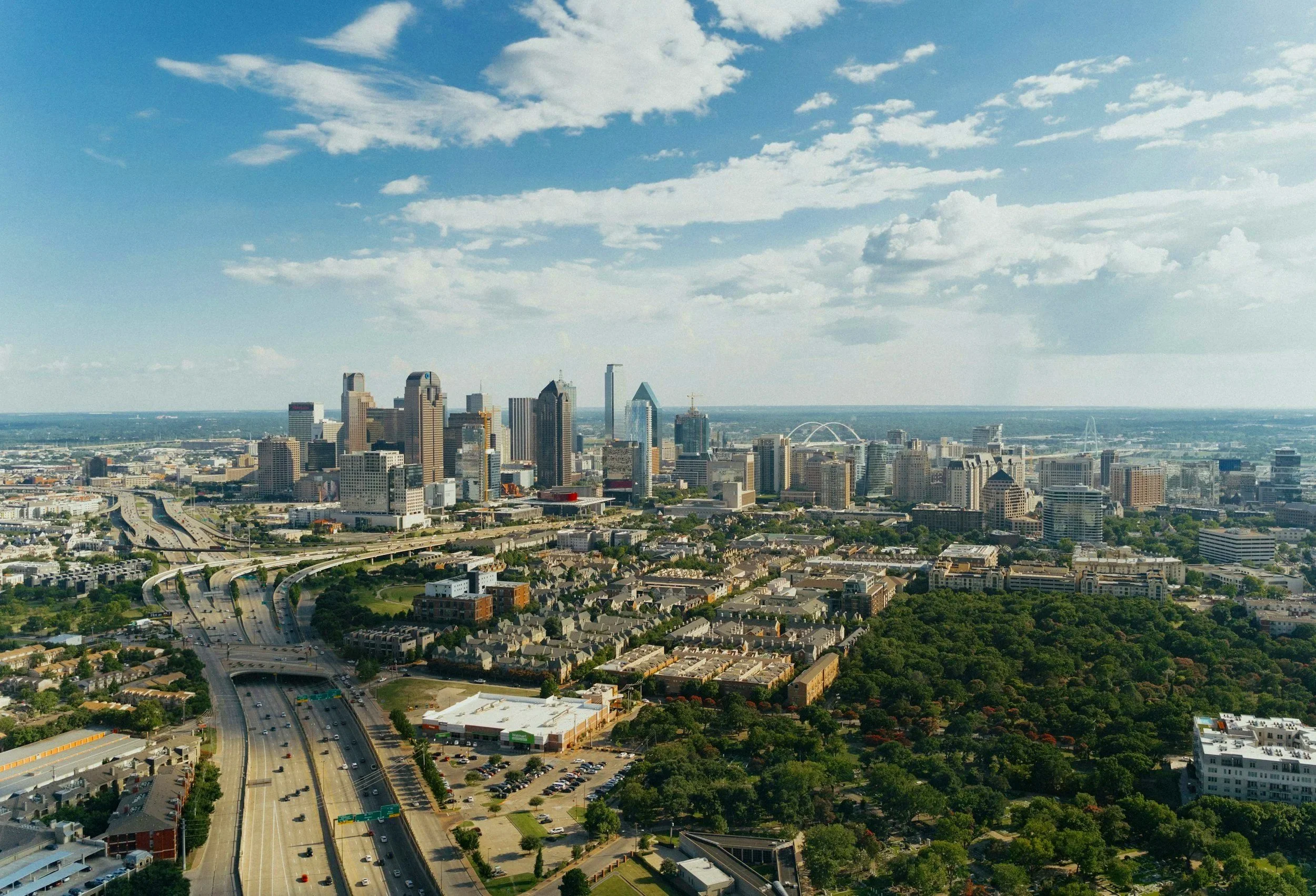 Aerial view of downtown Dallas, Texas with skyscrapers, highways, and green parks under a partly cloudy sky.