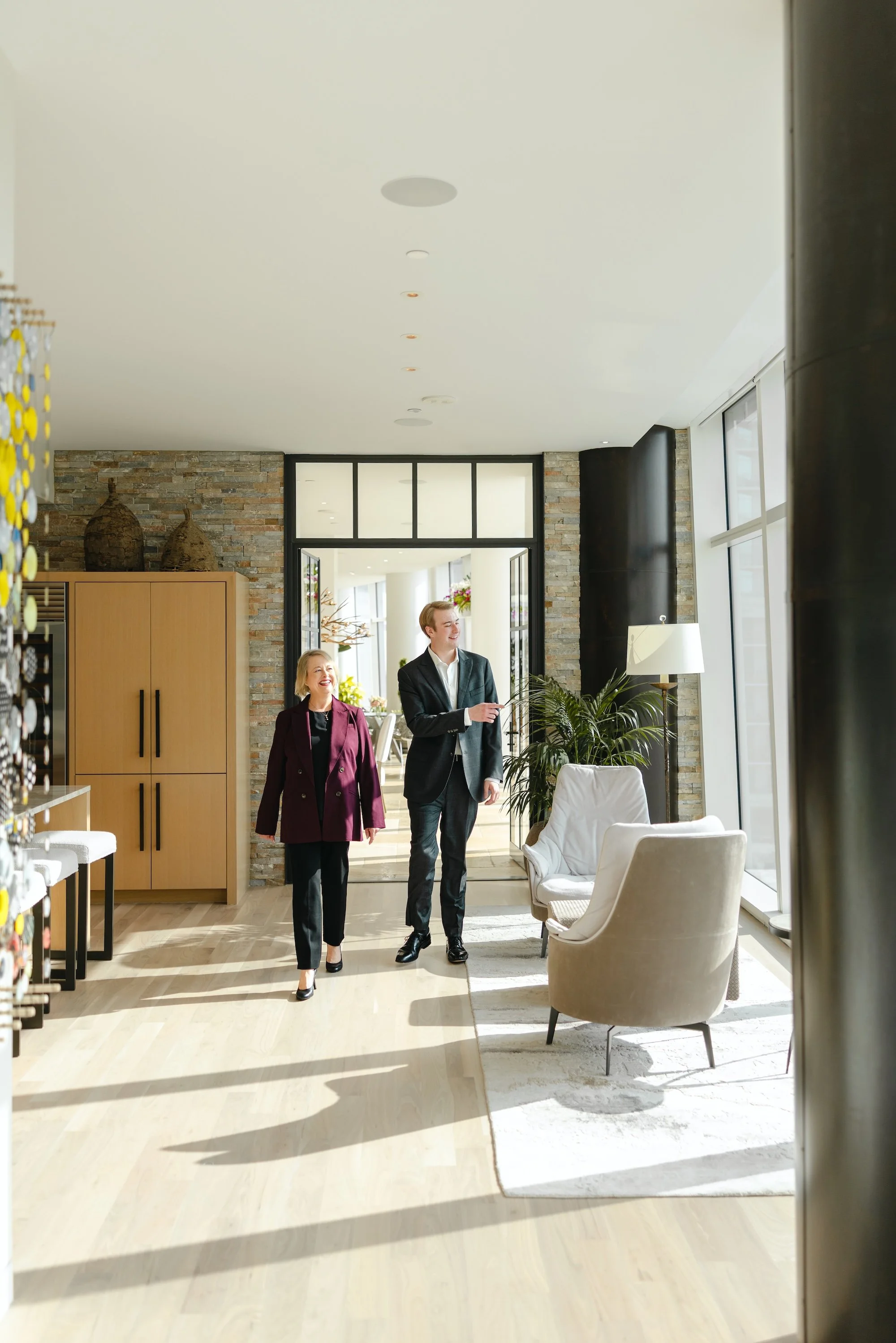 Two people, a man and a woman, walking inside a modern, well-lit room with large windows, a beige rug, and comfortable seating, smiling and engaging in conversation.