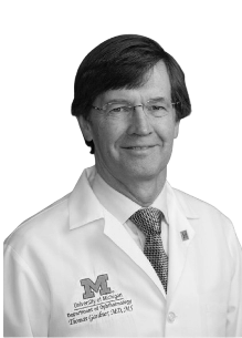 Black and white photo of a man in a white medical coat with a University of Michigan logo, wearing glasses and a tie, smiling.