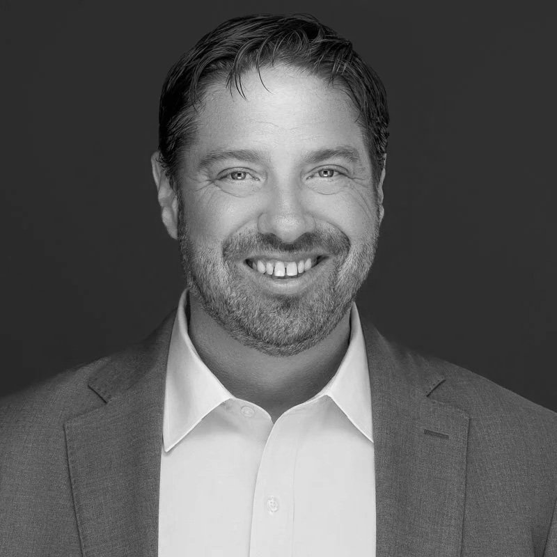 Black and white portrait of a smiling man with short hair and a beard, wearing a suit jacket and white shirt against a dark background.