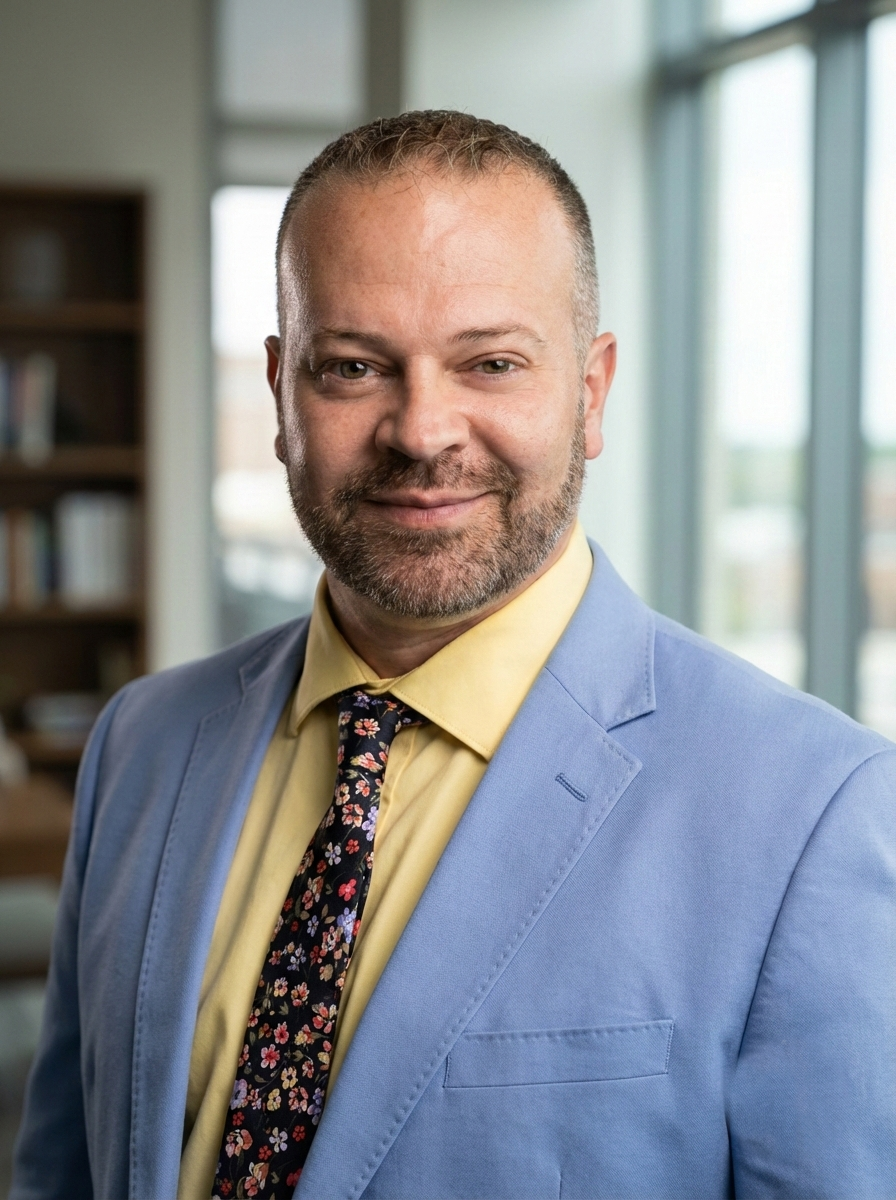 A man with a beard smiling, wearing a yellow shirt, a floral tie, and a light blue blazer, in an office with large windows and bookshelves in the background.
