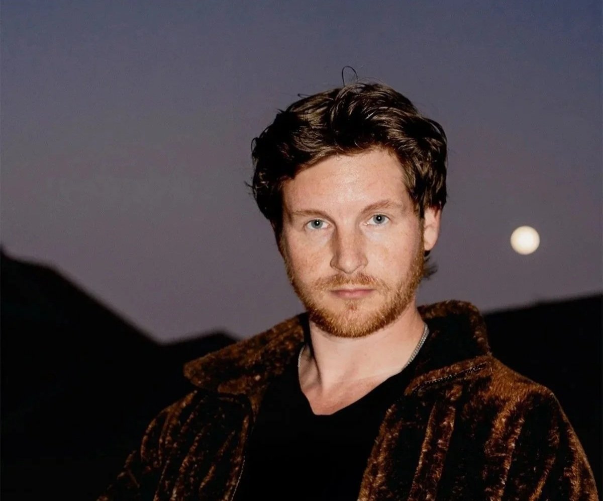 Portrait of a man with short brown hair and a beard, wearing a dark patterned jacket, against a twilight sky with a visible moon in the background.
