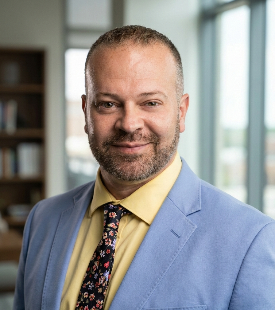 A man with short hair and a beard, dressed in a yellow shirt, floral tie, and light blue blazer, smiling in an office with large windows and bookshelves.