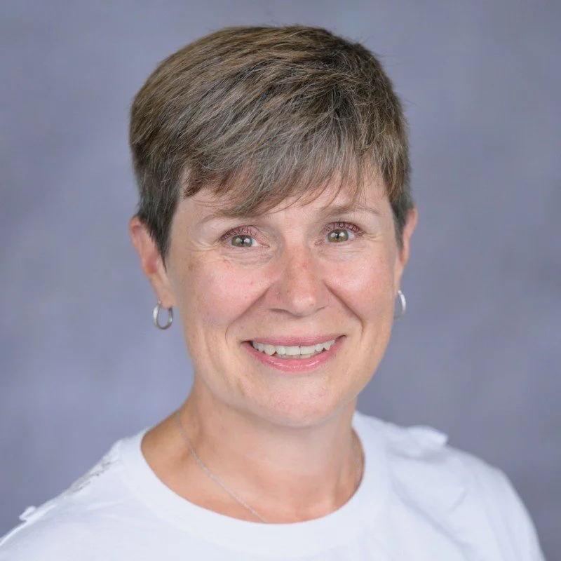 A smiling woman with short brown hair, wearing a white top and small hoop earrings, against a neutral background.