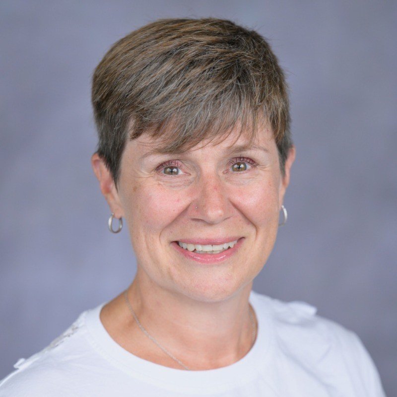 Close-up portrait of a middle-aged woman with short brown hair, wearing a white top and silver hoop earrings, smiling against a neutral background.