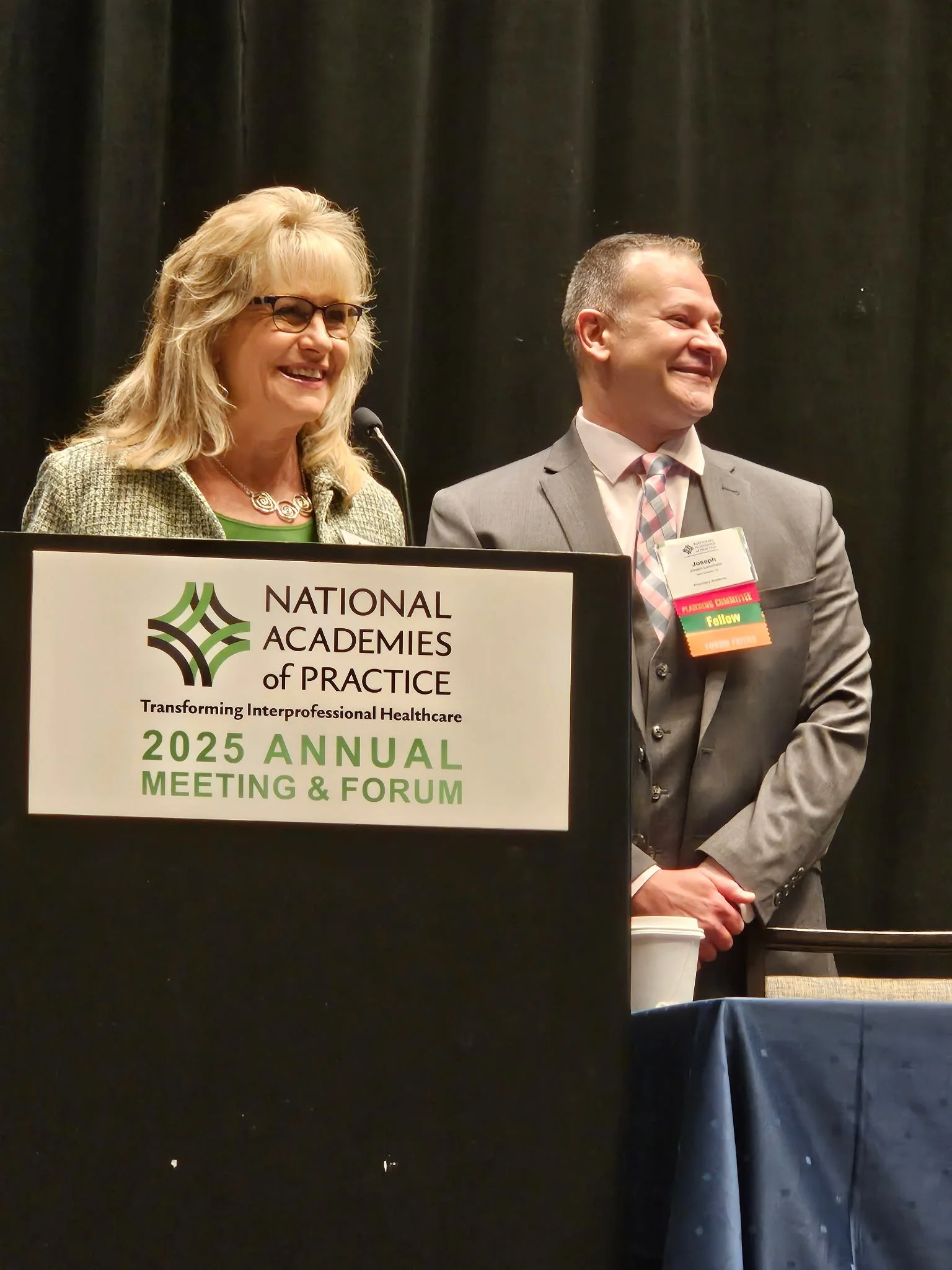 Two people smiling and standing behind a podium at the 2025 Annual Meeting & Forum of the National Academies of Practice, with a woman on the left and the CEO on the right.