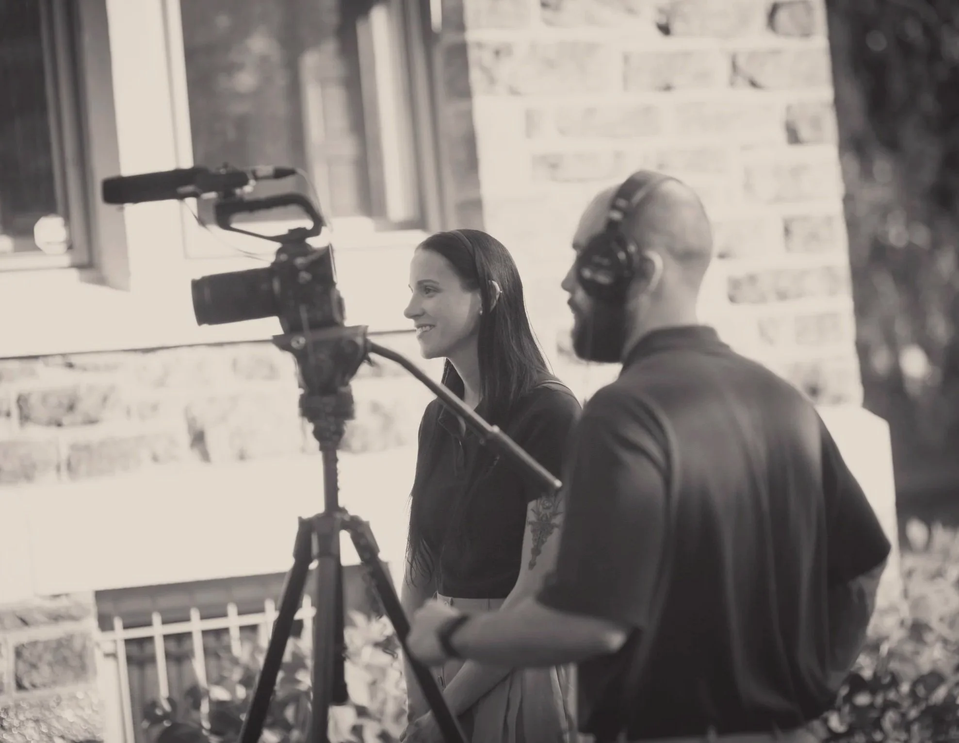 Two people, a woman and a man, sitting outdoors with a camera on a tripod, possibly making a video or film, both wearing headphones, with a brick wall and window in the background.