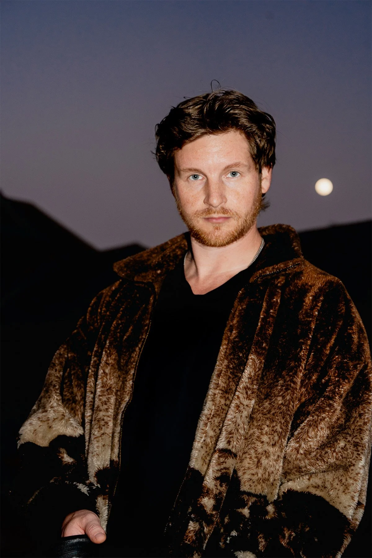 A man with light skin, red hair, and a beard, wearing a brown patterned fur coat over a black shirt, standing outdoors during dusk with a dark sky and the moon in the background.