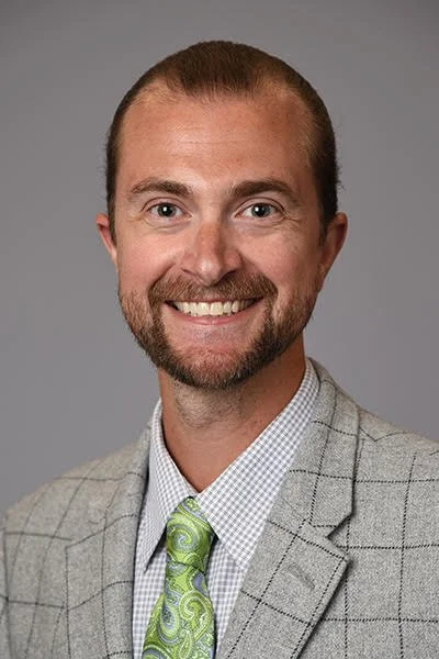 Portrait of a smiling man with short light brown hair, a beard, wearing a gray plaid blazer, a white checkered shirt, and a light green paisley tie, against a gray background.