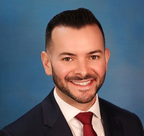 A professional portrait of a man smiling, dressed in a navy suit, white shirt, and red tie, with a blue background.