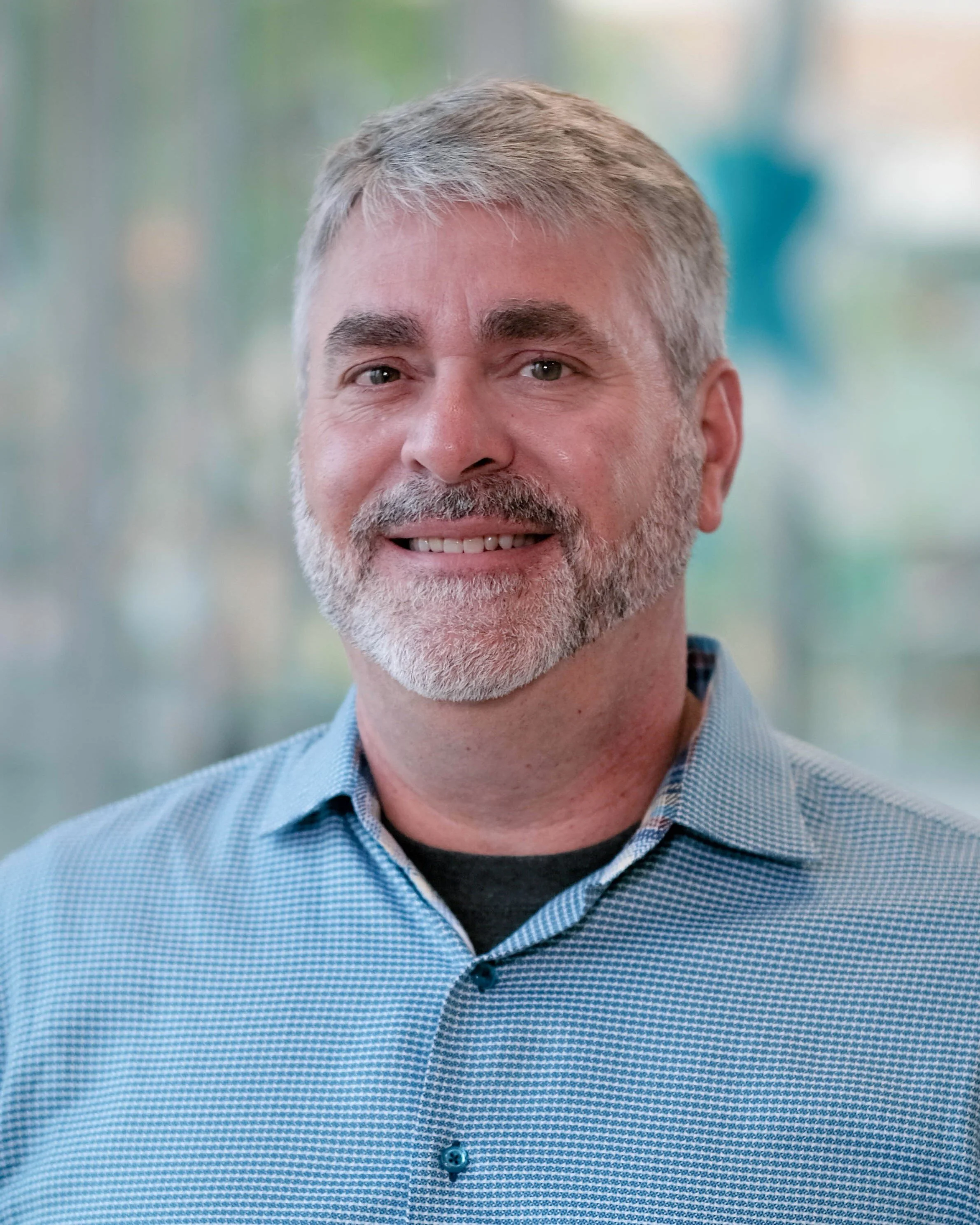 Close-up portrait of a middle-aged man with gray hair and a beard, smiling, wearing a blue checkered shirt, in an indoor setting with a softly blurred background.