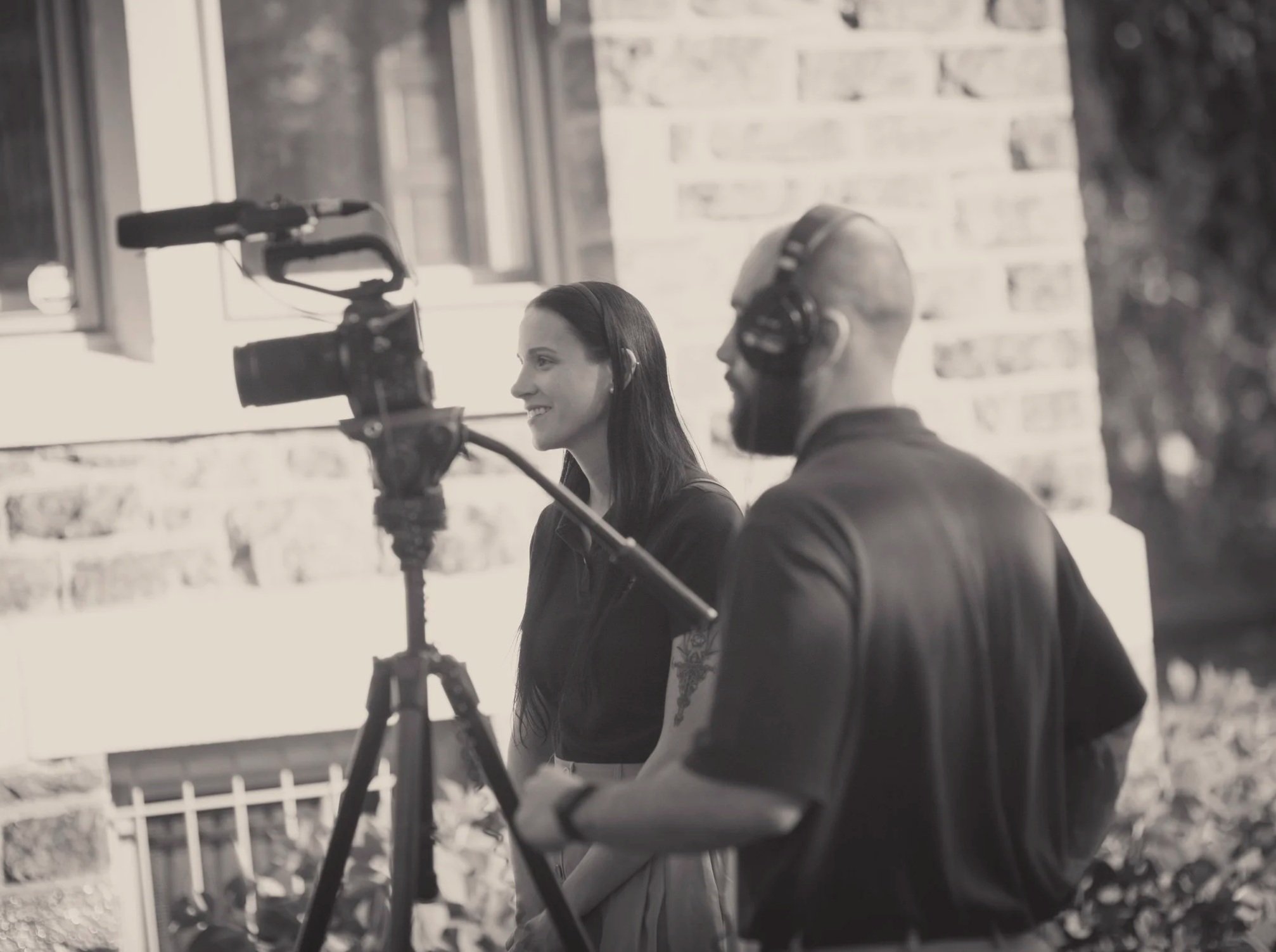 A woman and a man working on a video shoot with a camera on a tripod, woman smiling, man wearing headphones, both focusing on filming outdoors near a house.