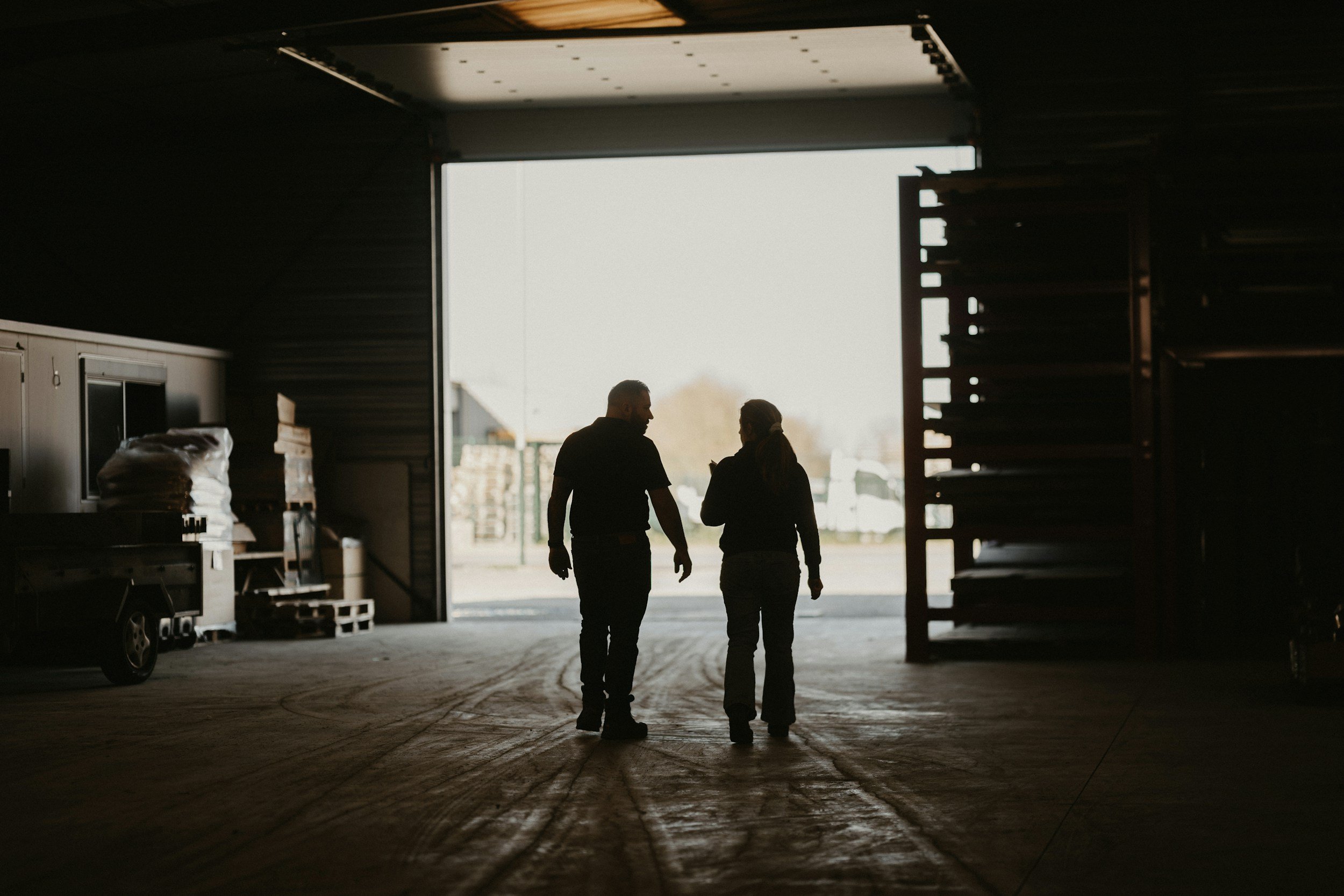 Silhouettes of a man and woman walking and talking inside a warehouse or storage area with an open bay door, revealing outdoor storage containers and machinery.