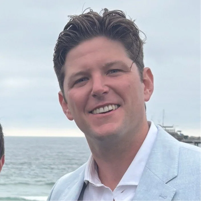 A smiling man with brown hair wearing a light gray blazer over a white shirt, outdoors by the water with an overcast sky.