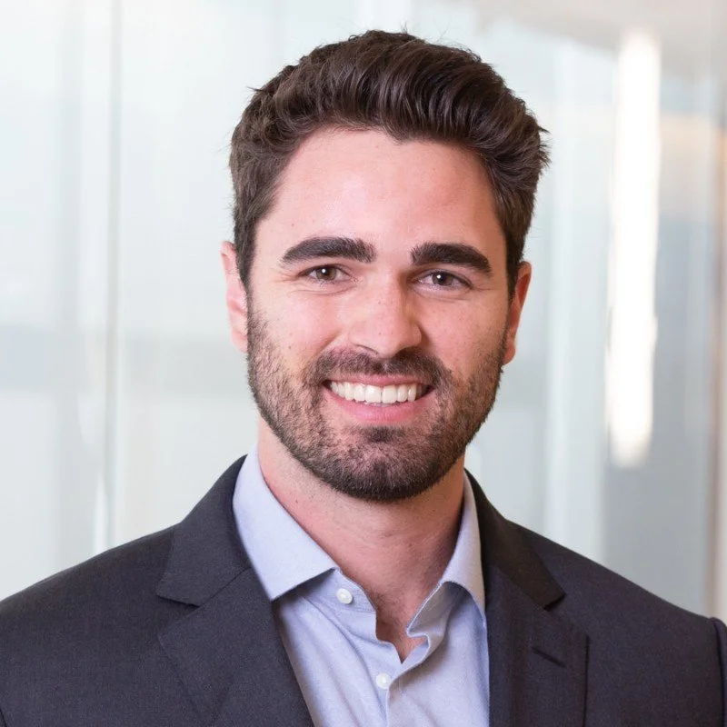 A man with brown hair, beard, and blue eyes wearing a dark suit and light blue shirt, smiling in an office setting with blurred glass walls in the background.