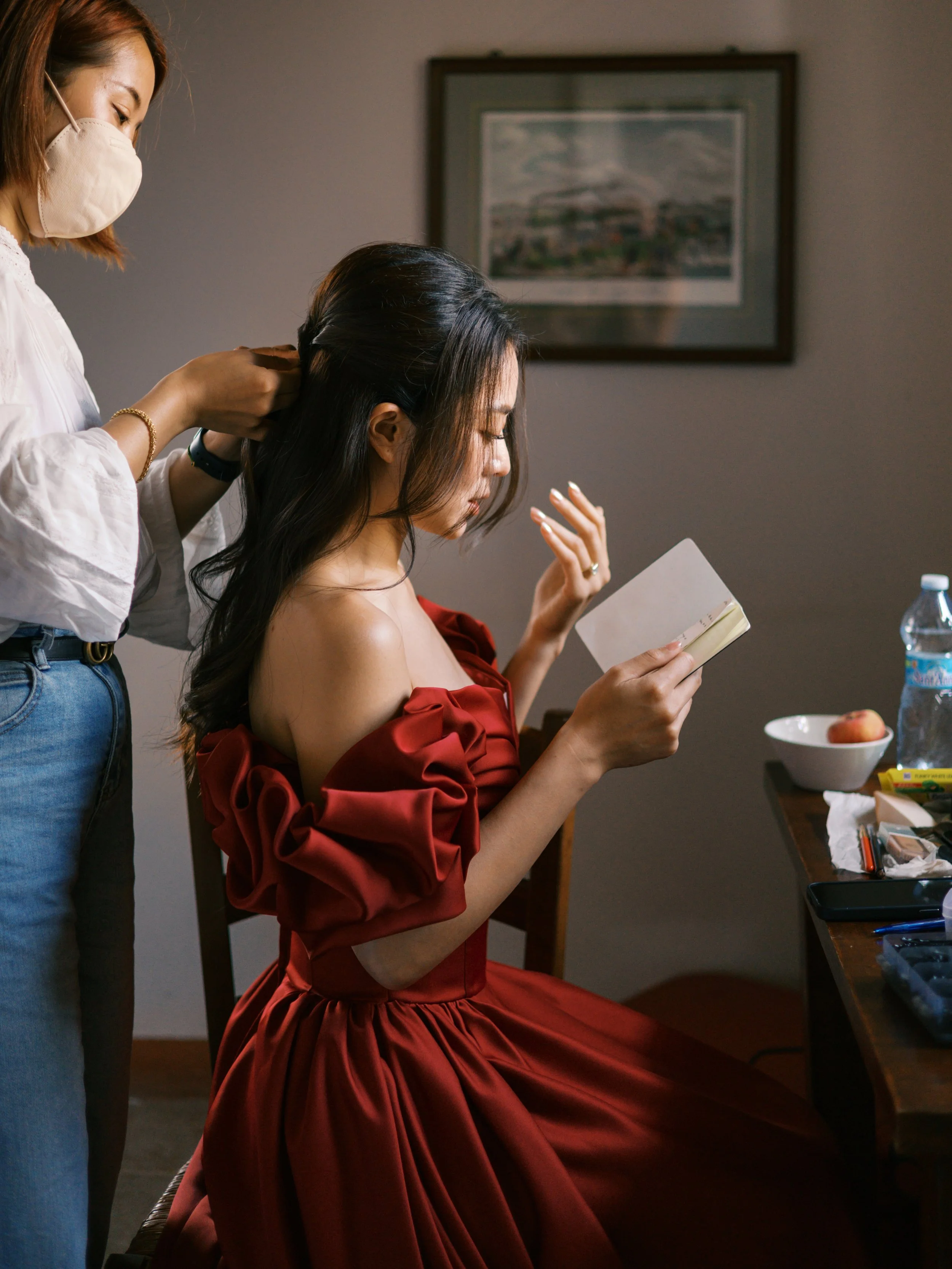 A woman in a red dress reading a card while a hairstylist styles her hair in a dimly lit room.