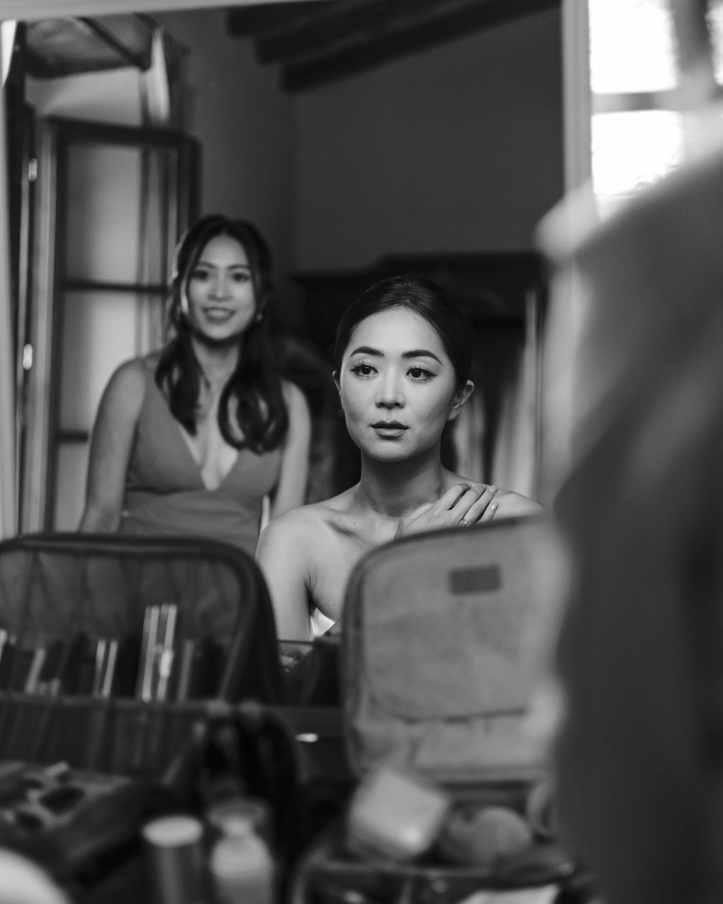 Two women in a dressing room, one sitting in front of a mirror and the other standing behind her. The woman in front is looking at her reflection, while the woman behind is smiling and looking toward the camera. Makeup and beauty products are on the table in front of them.