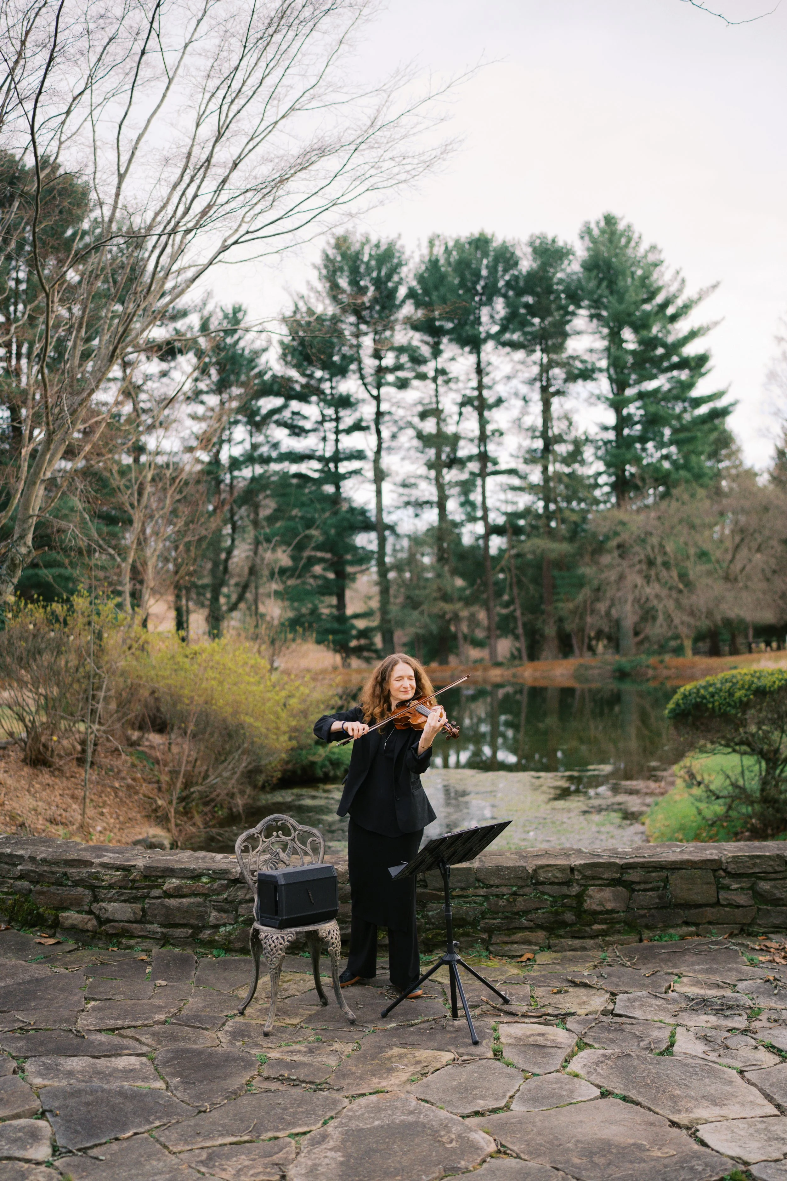 A woman in black clothing playing the violin outdoors near a pond surrounded by trees.