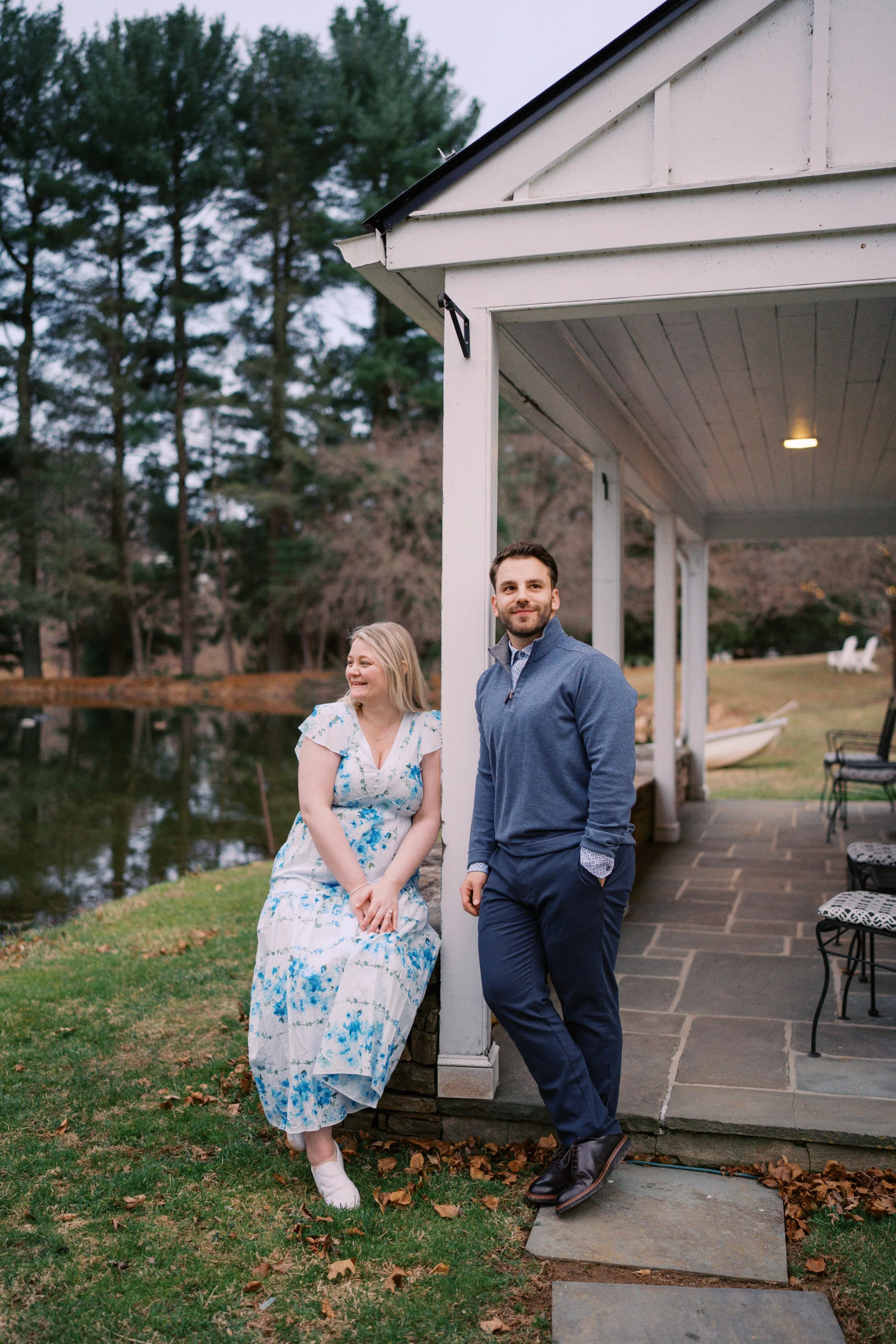 A woman in a floral dress sitting by a body of water, smiling and looking at a man in a blue jacket and dark pants, standing near a white porch, in an outdoor setting with trees and chairs.