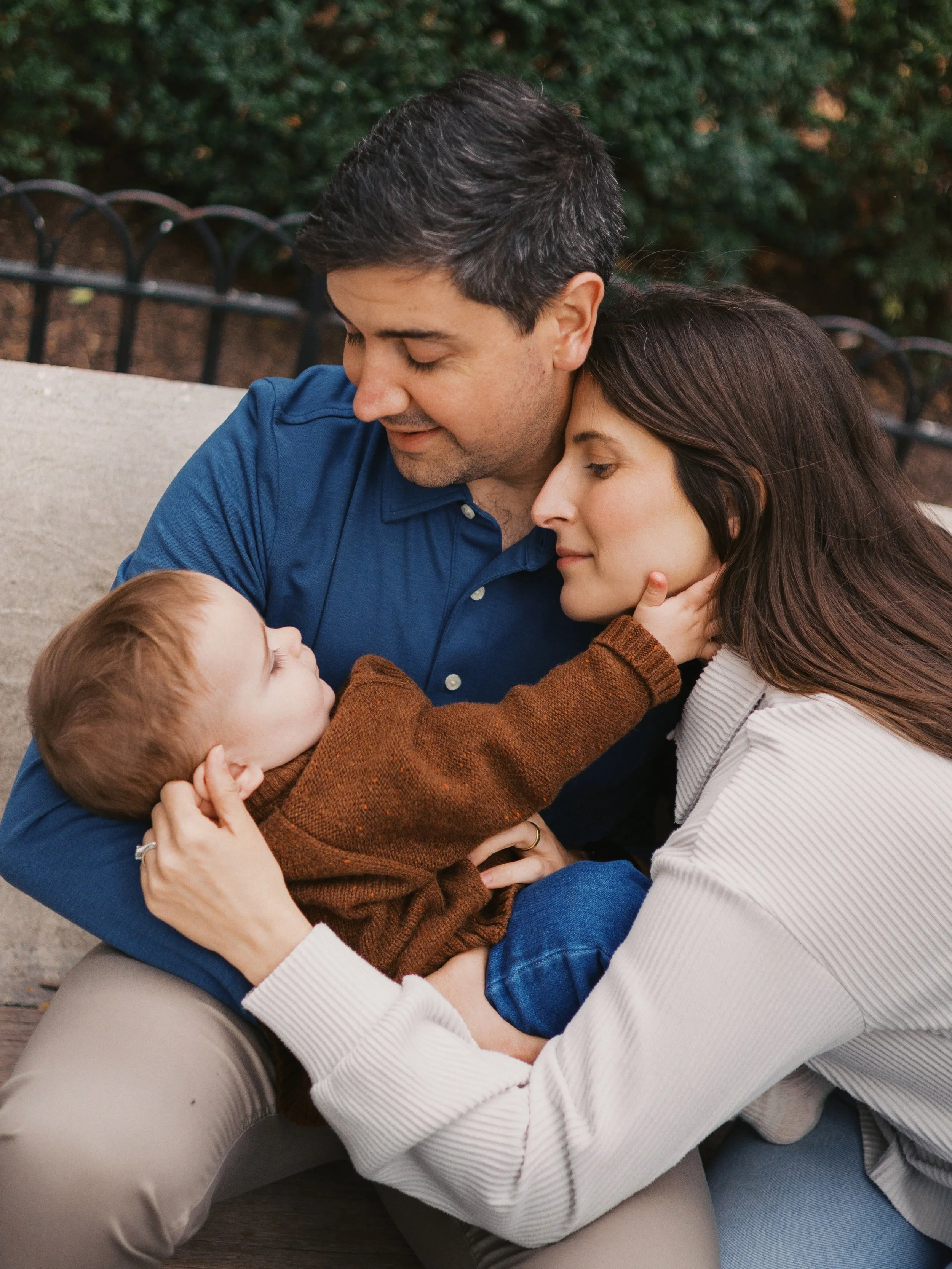 A family of three: a father, mother, and young son sharing a tender moment outdoors on a park bench, with the son touching the mother's face.