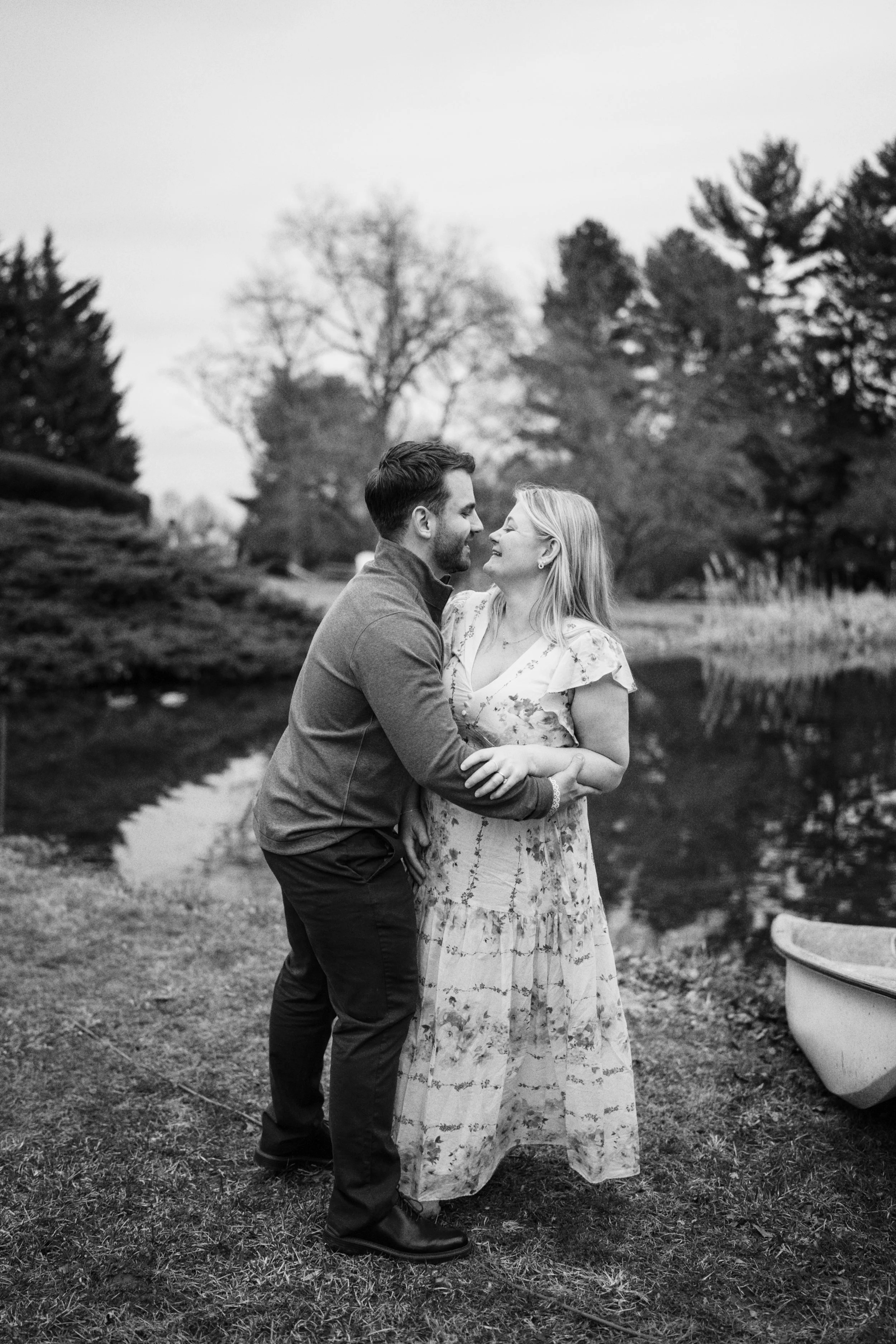 A black and white photo of a couple smiling and embracing near a pond in a park. The man is wearing a jacket and the woman is in a floral dress with ruffles.
