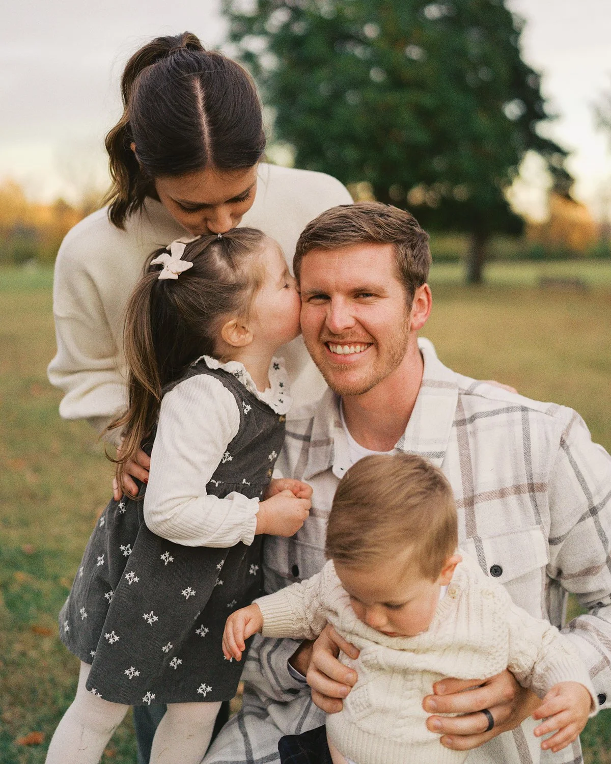 A family of four in Chester County, PA, a man, woman, and two young children outdoors in a park, with the girl giving the man a kiss on the cheek and the woman kissing the girl's head, during fall.