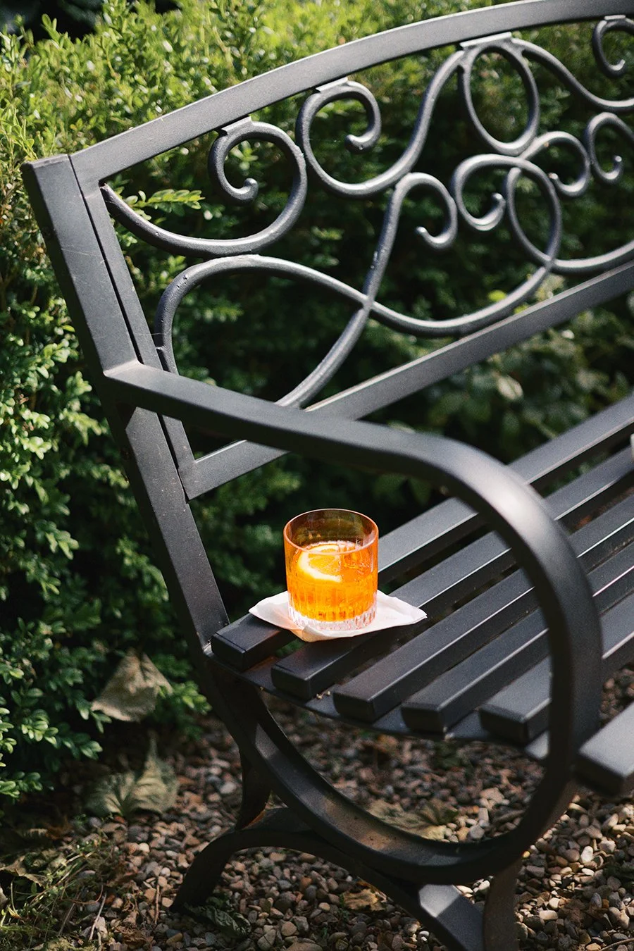 A black metal park bench with ornate lattice armrests, placed on a gravel surface with green bushes in the background. On the bench, there is a glass of orange-colored beverage with a lemon slice, resting on a white napkin.