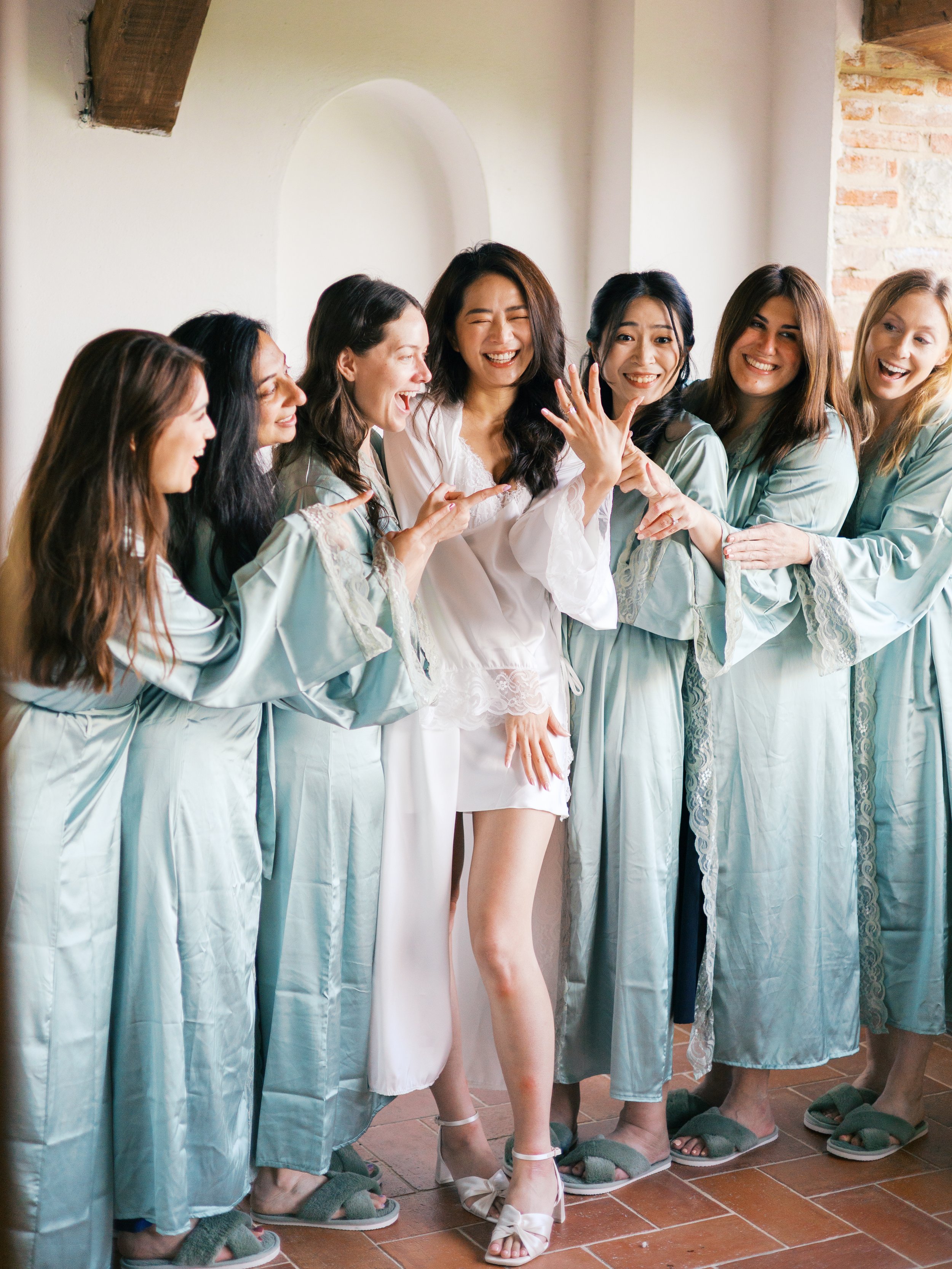 Group of women in matching pale blue robes and slippers, happily surrounding a bride in white satin pajamas, showing her engagement ring in a joyful celebration.