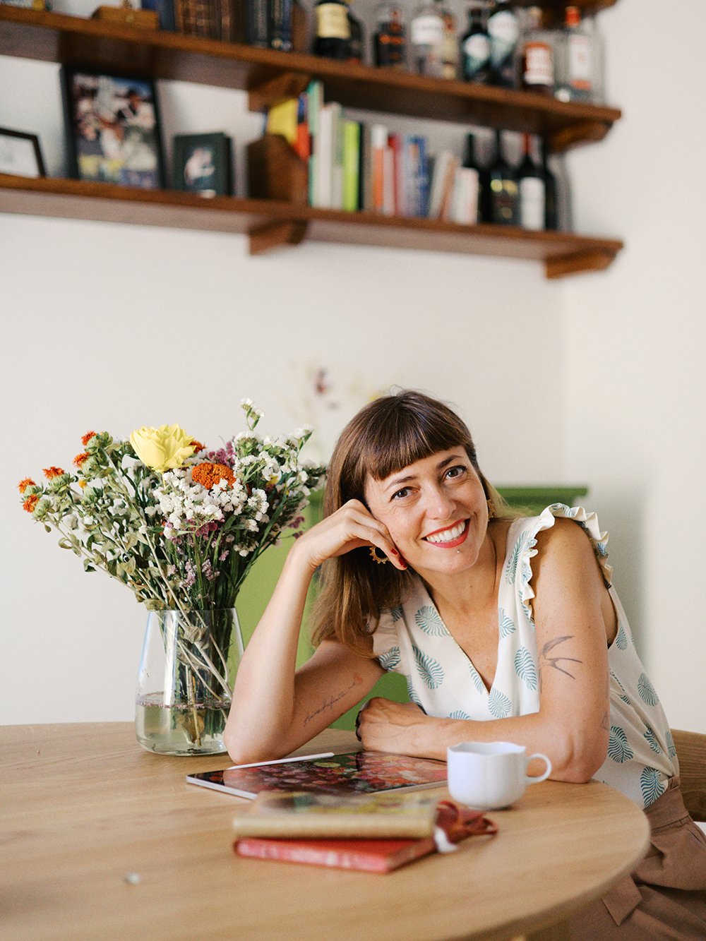 A woman with brown hair and tattoos on her arm sitting at a wooden table with a smile, supporting her head with her hand. There is a vase of mixed flowers, a ceramic cup, and some books on the table. In the background, wooden shelves with books and bottles.