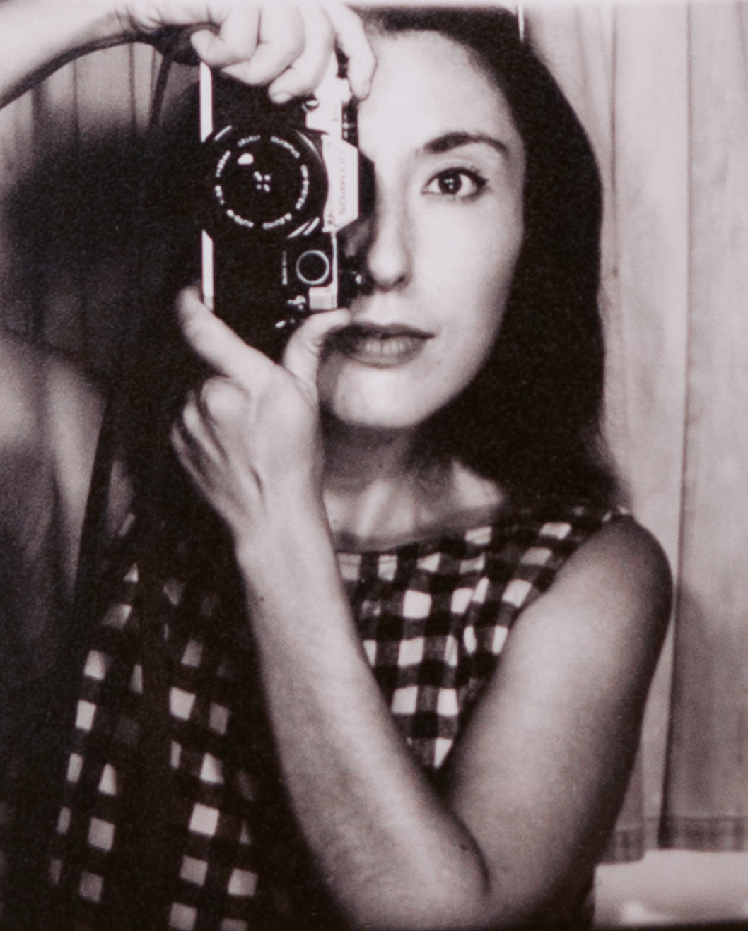 A woman taking a selfie in a mirror with a camera, wearing a sleeveless checkered top, with dark hair and light skin, in front of wooden paneling.