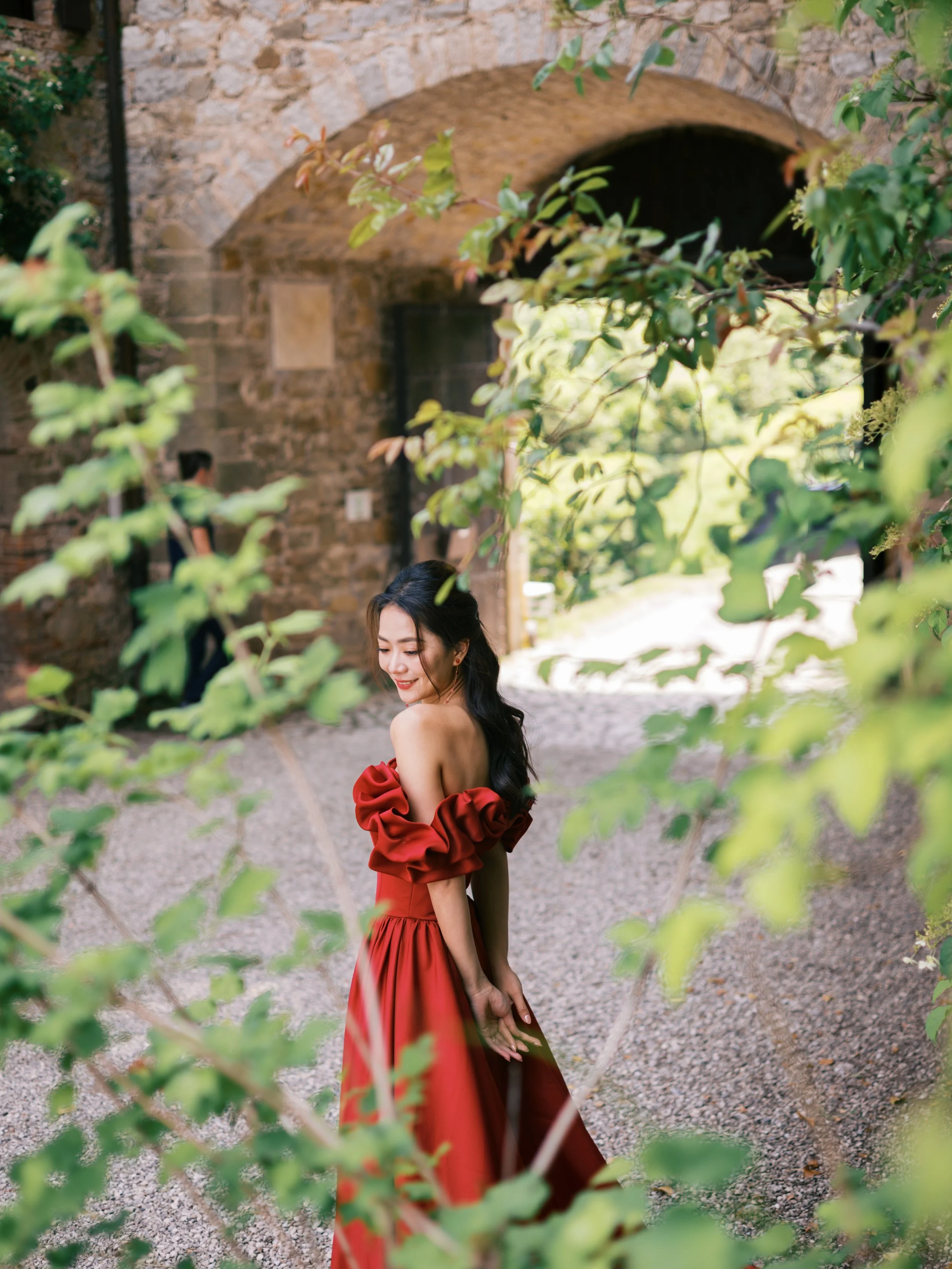 A woman in a red dress is standing outdoors near a stone archway, smiling and looking downward, with greenery in the foreground.