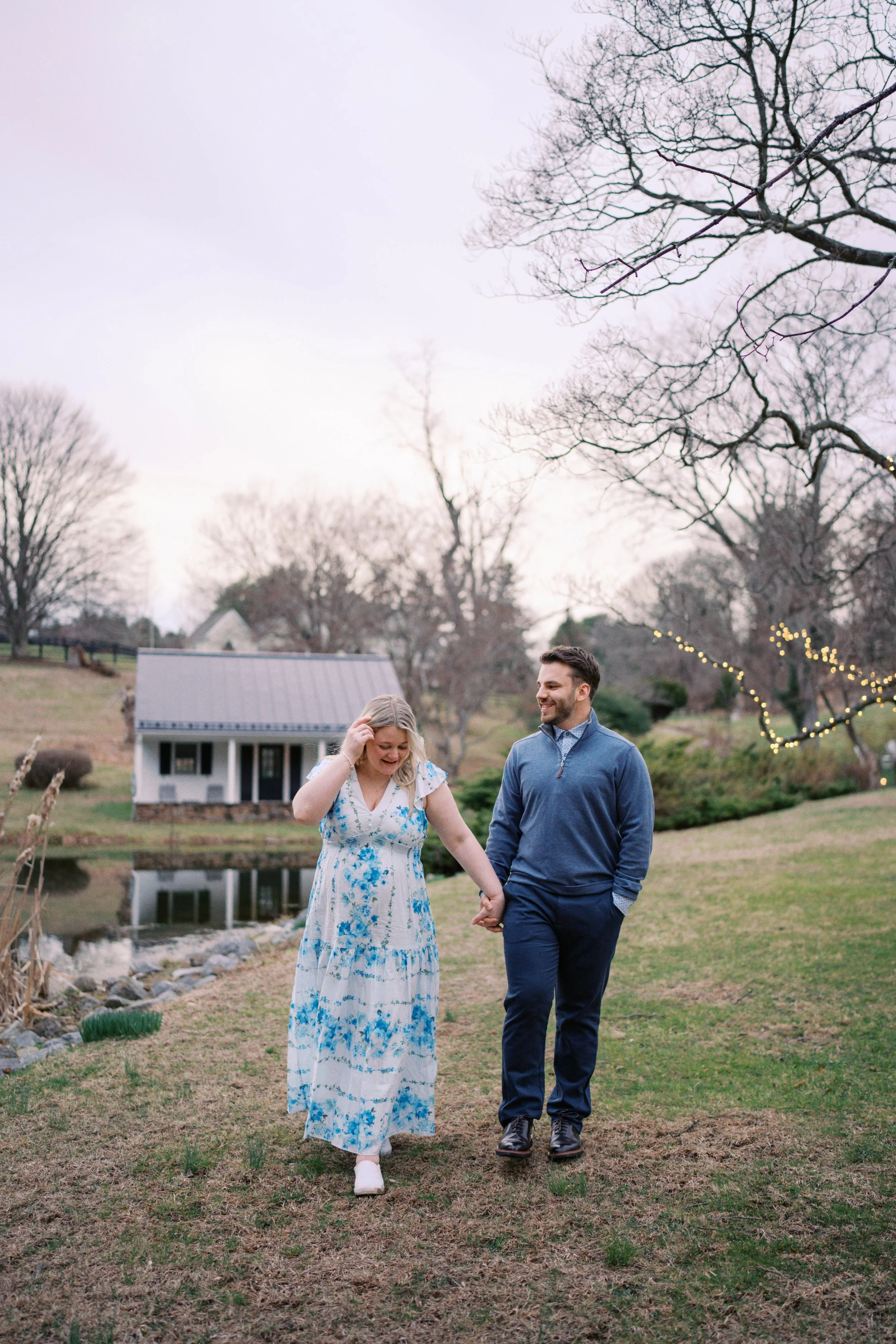 A couple walking hand in hand outdoors near a pond, with leafless trees and a small house in the background during dusk.