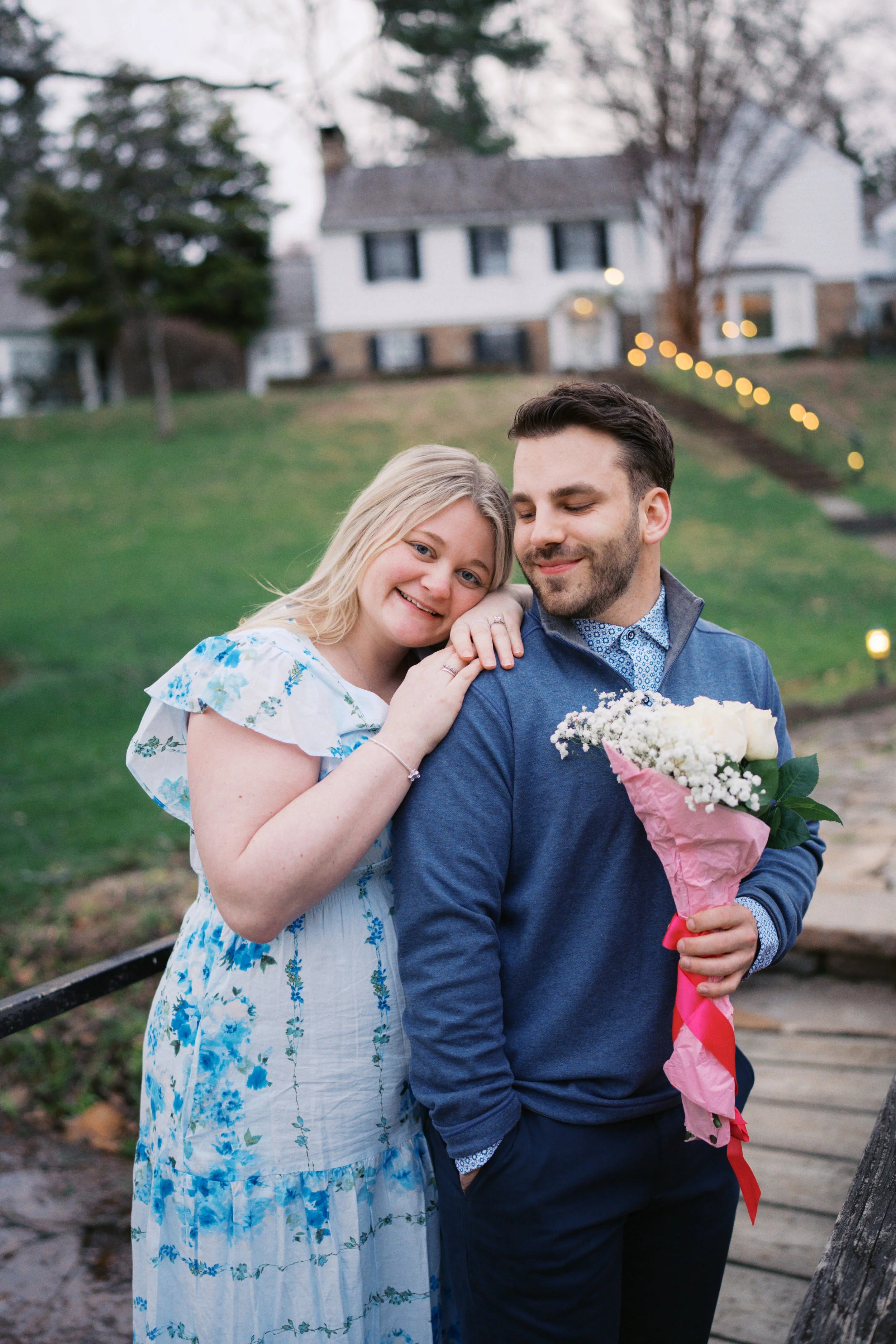 A happy couple stands close together outdoors on a bridge, with a woman resting her hand on a man's shoulder who is holding a bouquet of white flowers. A house and string lights are visible in the background.