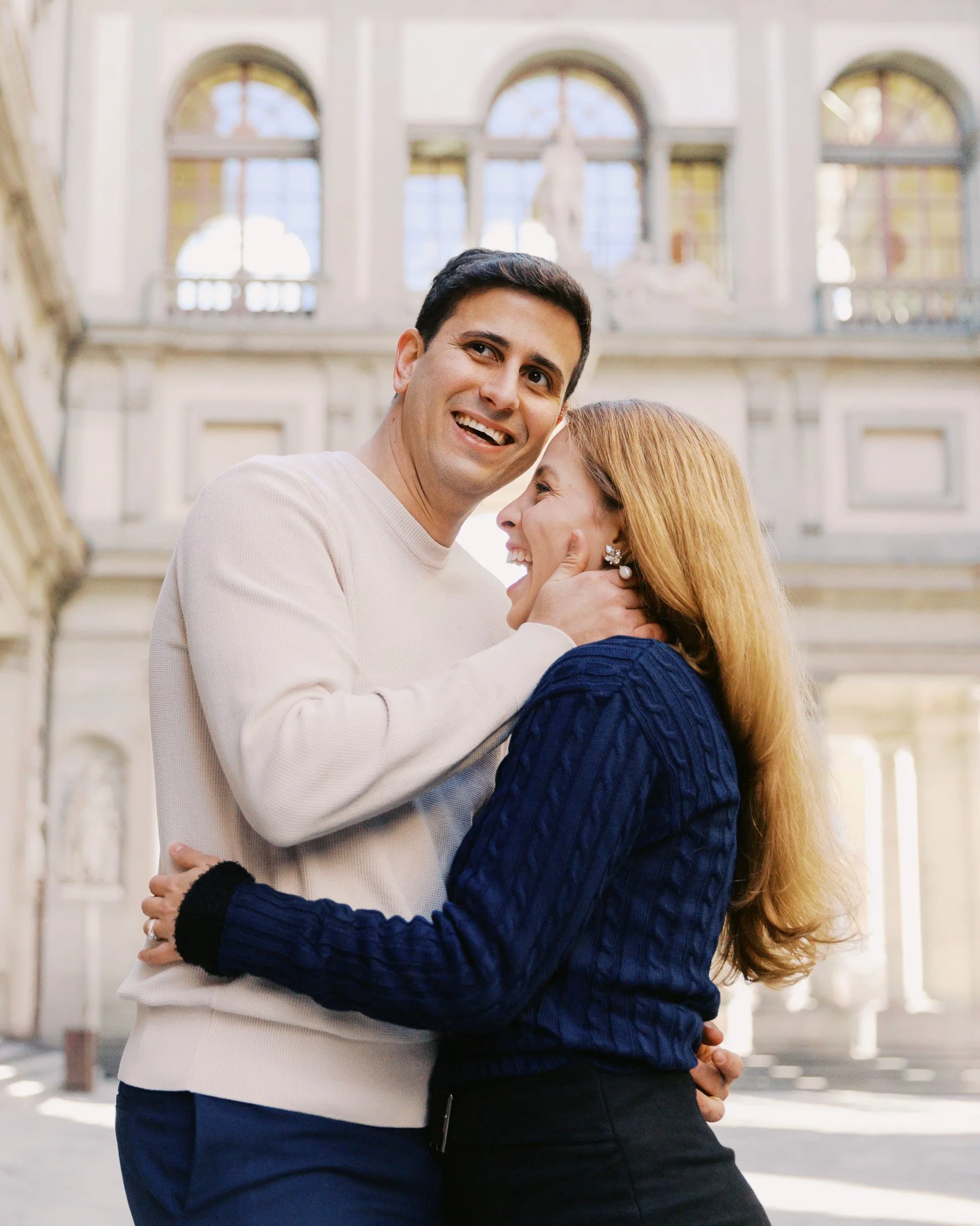 A smiling man and woman embracing each other near the Uffizi Gallery in Florence, Italy with large arched windows and classical statues in the background.