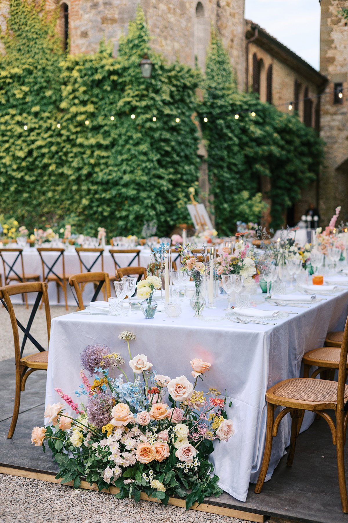 Outdoor elegantly decorated dining table with floral centerpieces, candles, and glassware, set against a backdrop of a stone building covered in green ivy during dusk.