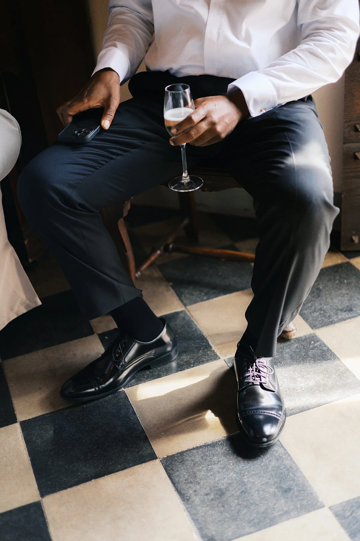 Close-up of a man in formal attire holding a wine glass and a smartphone, sitting on a wooden chair with black and white checkered floor tiles.