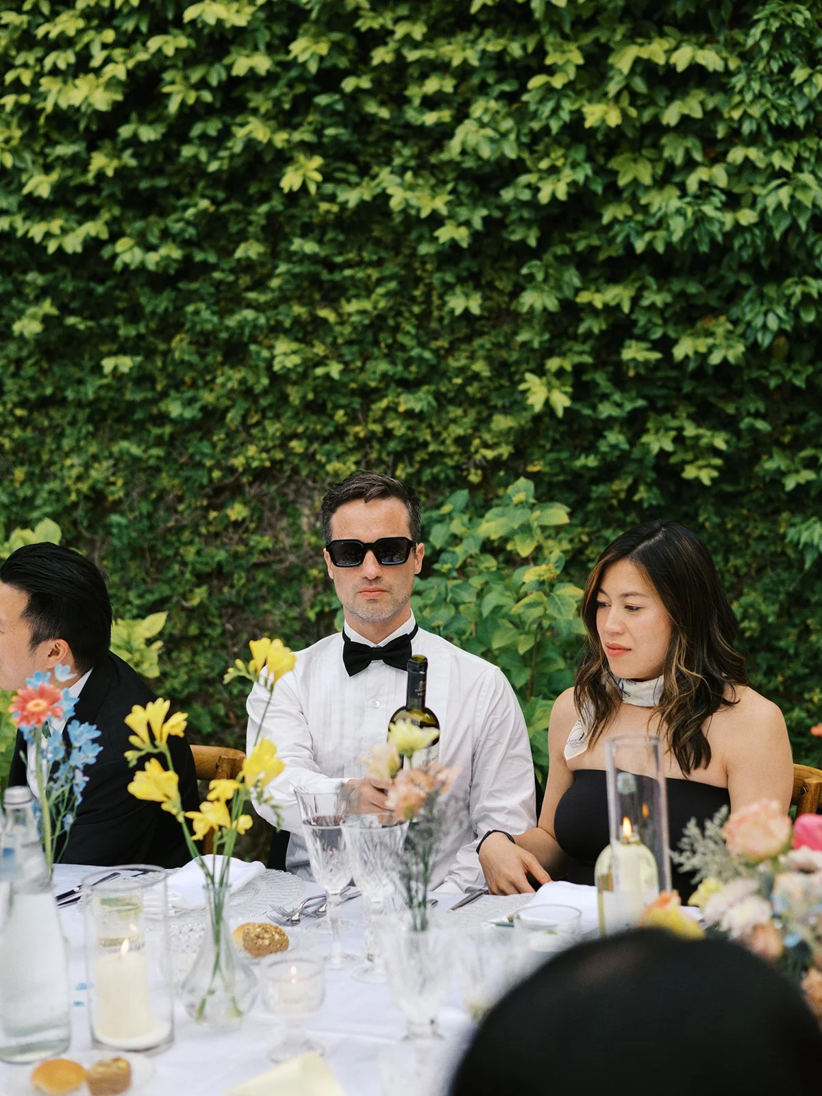 People sitting at a decorated outdoor table, with greenery in the background, including a man in sunglasses and a woman in a black dress.