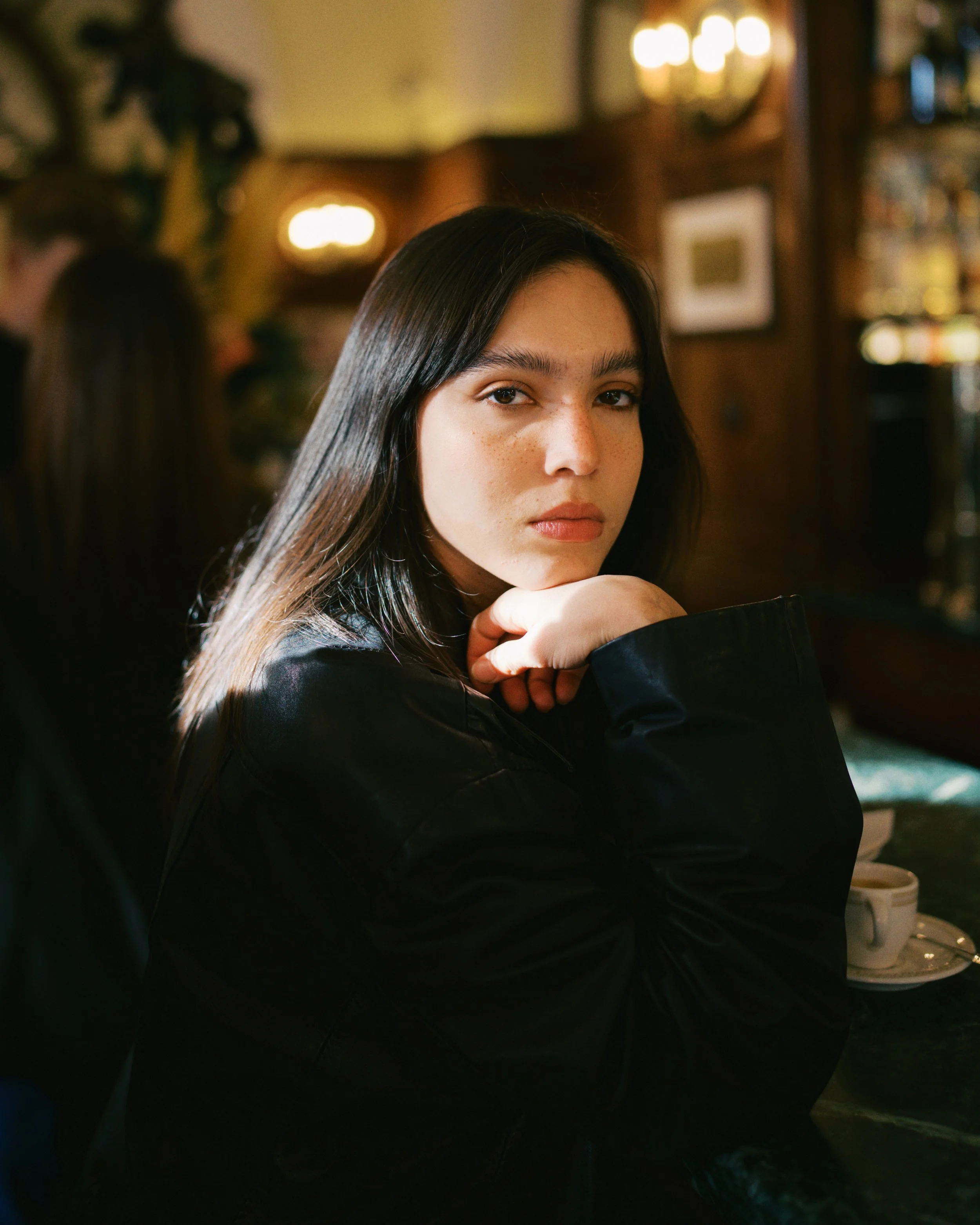 A young woman in Florence, Italy with dark hair and freckles sitting at a bar counter, resting her chin on her hand, gazing at the camera, in a dimly lit pub or restaurant.